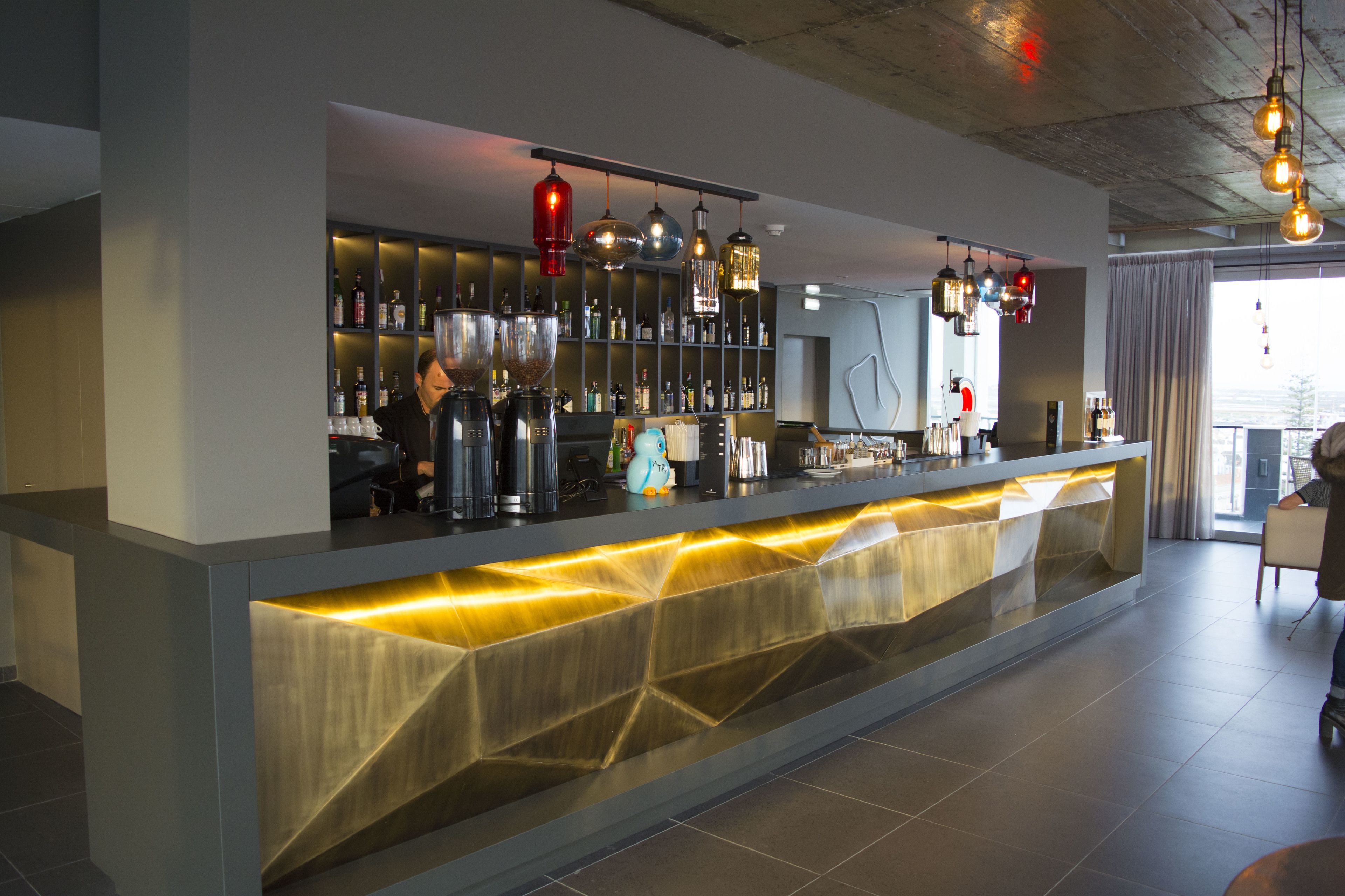 A modern bar area with a counter, shelves of bottles, and hanging lights.