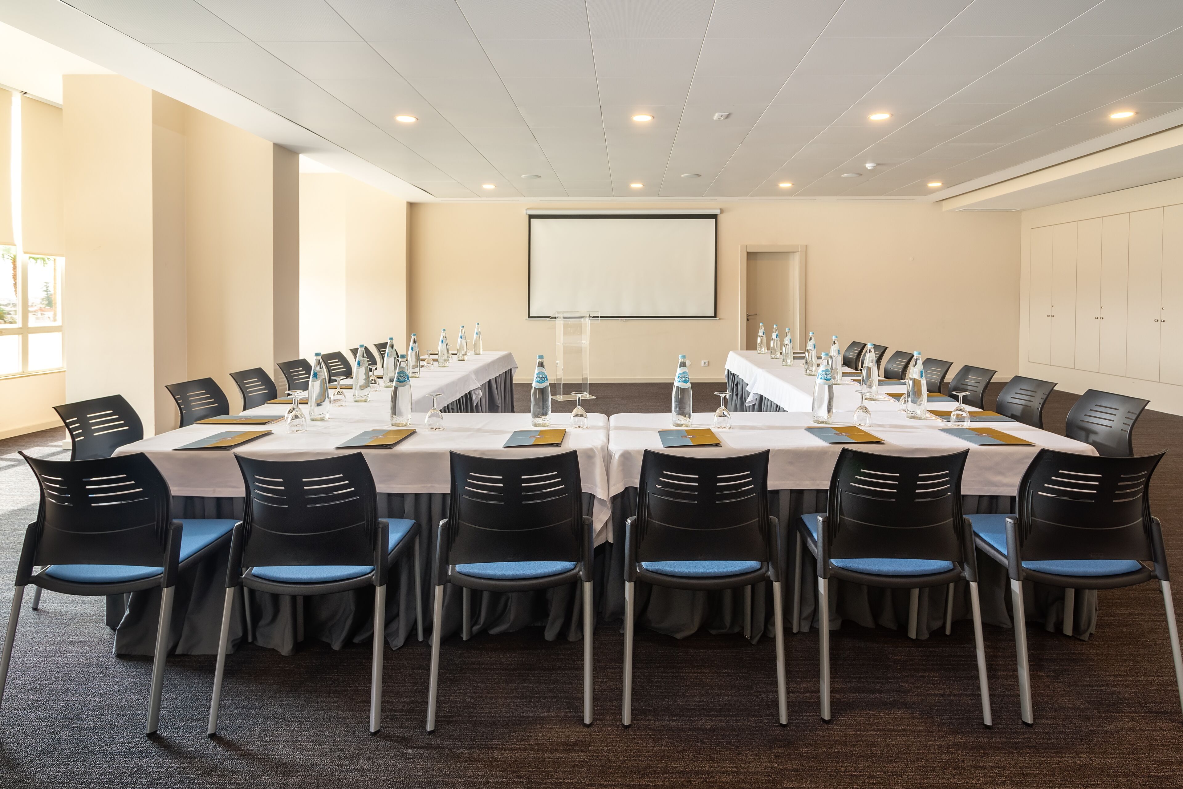 A conference room with a long table, chairs, water bottles, and a projector screen.