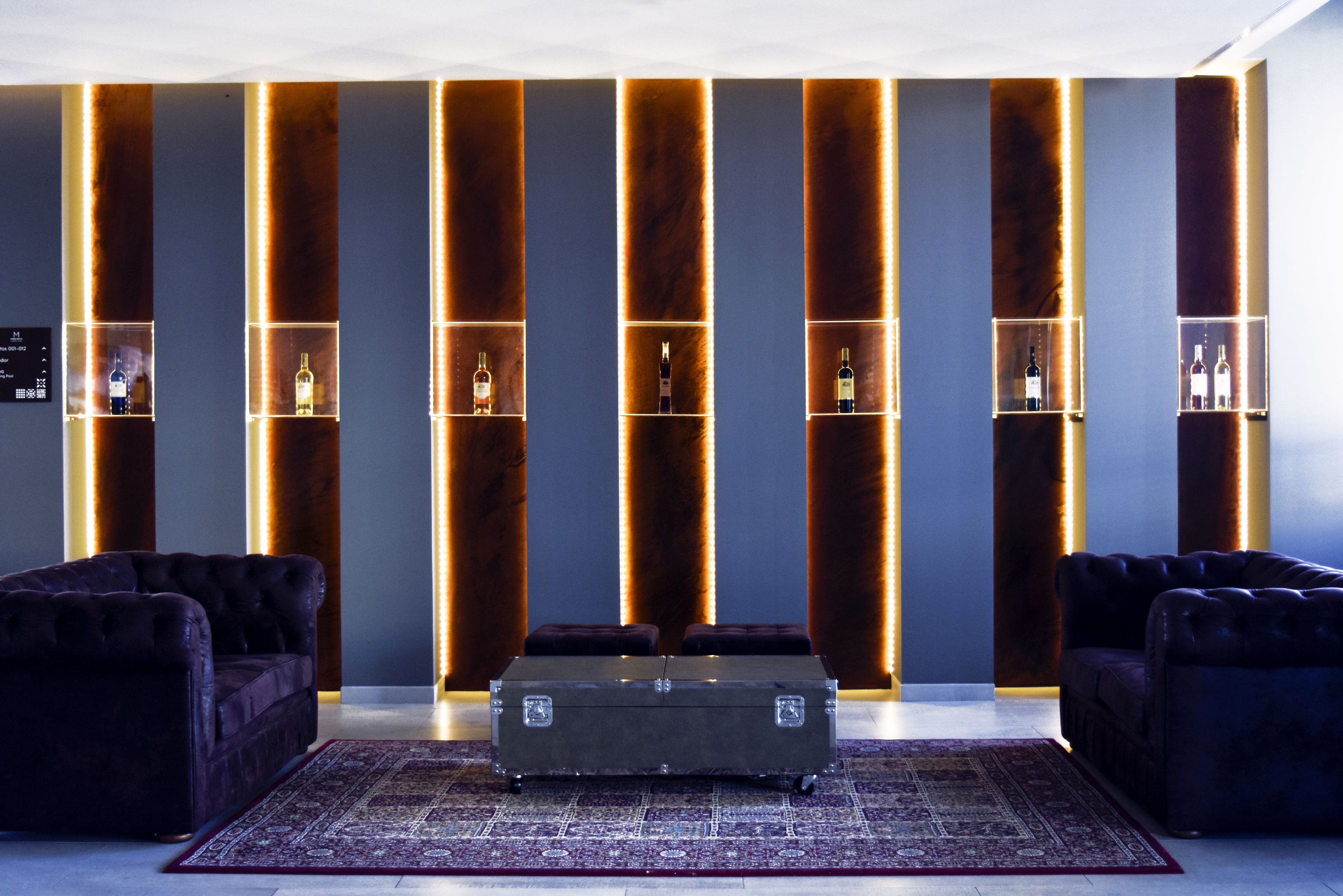 Un hall d’entrée moderne, avec un ensemble de canapés aux couleurs sombres, un pouf central et un mur doté d’étagères éclairées où sont exposées des bouteilles.