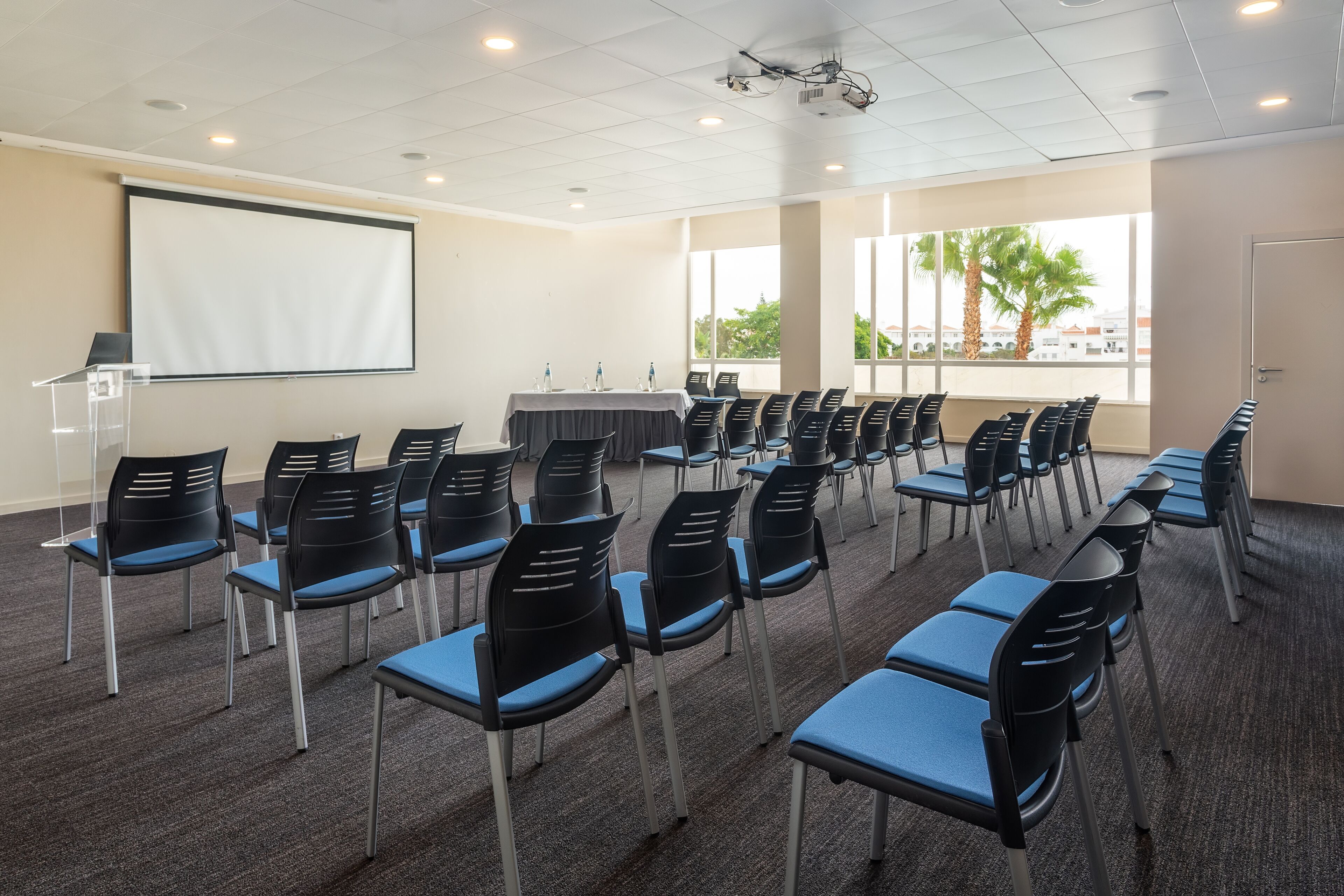 A conference room with rows of chairs, a podium, a projector screen, and a table with water bottles.