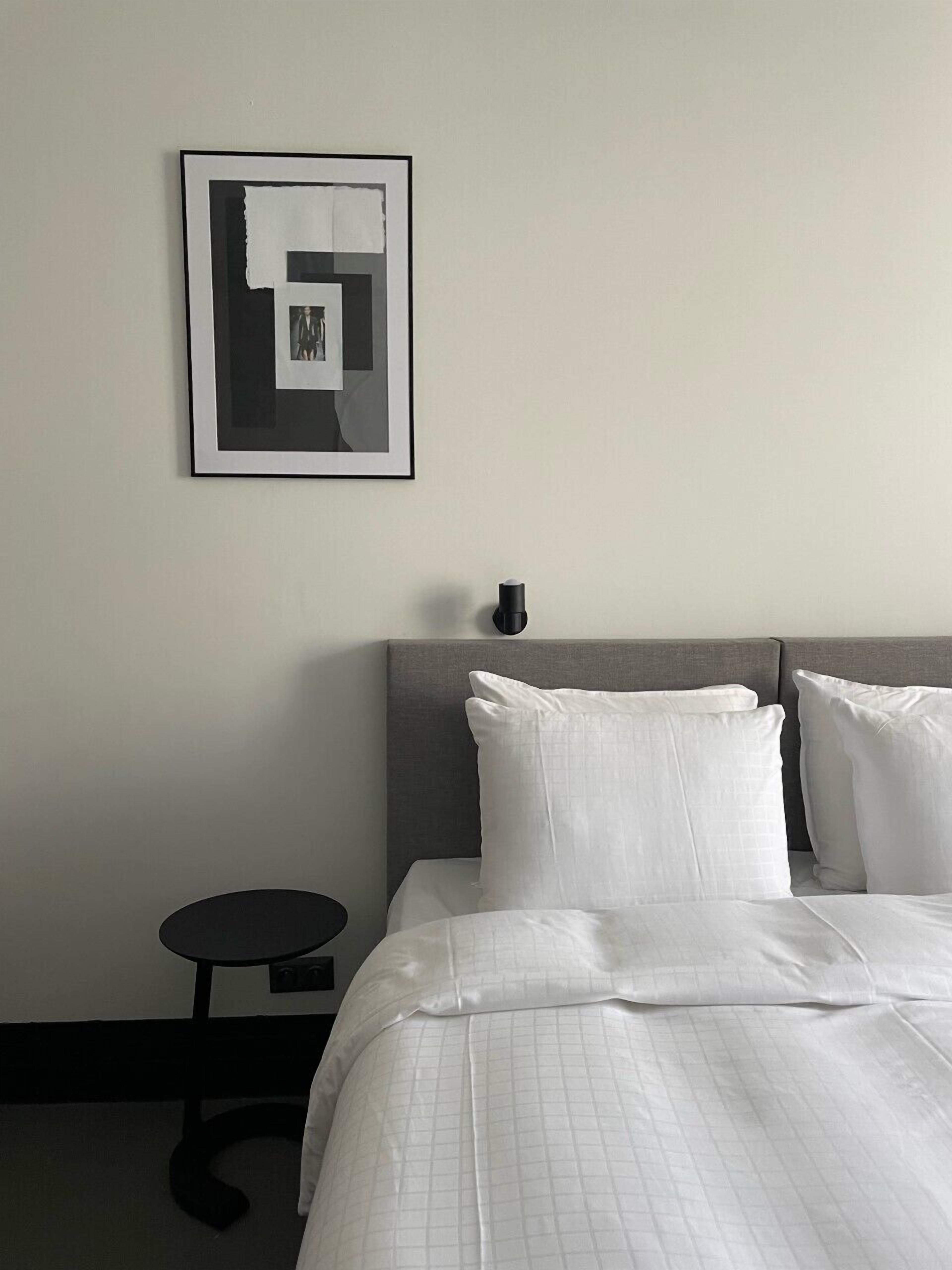 A hotel room with a bed, pillows, a framed artwork on the wall, and a small table.