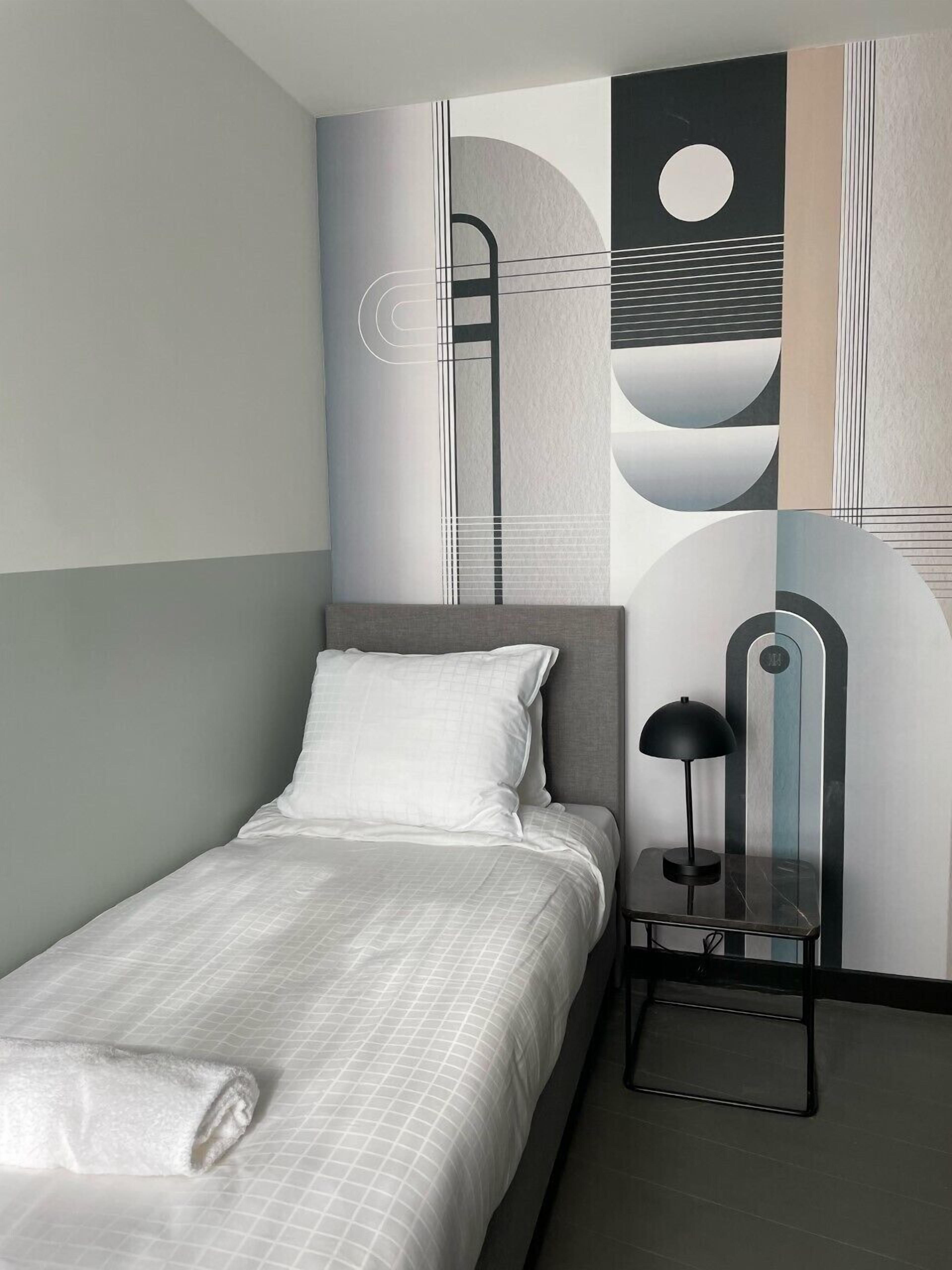 Een net opgemaakt bed met een wit kussen en een opgerolde handdoek, een zwarte bureaulamp en een muur met abstracte geometrische patronen.