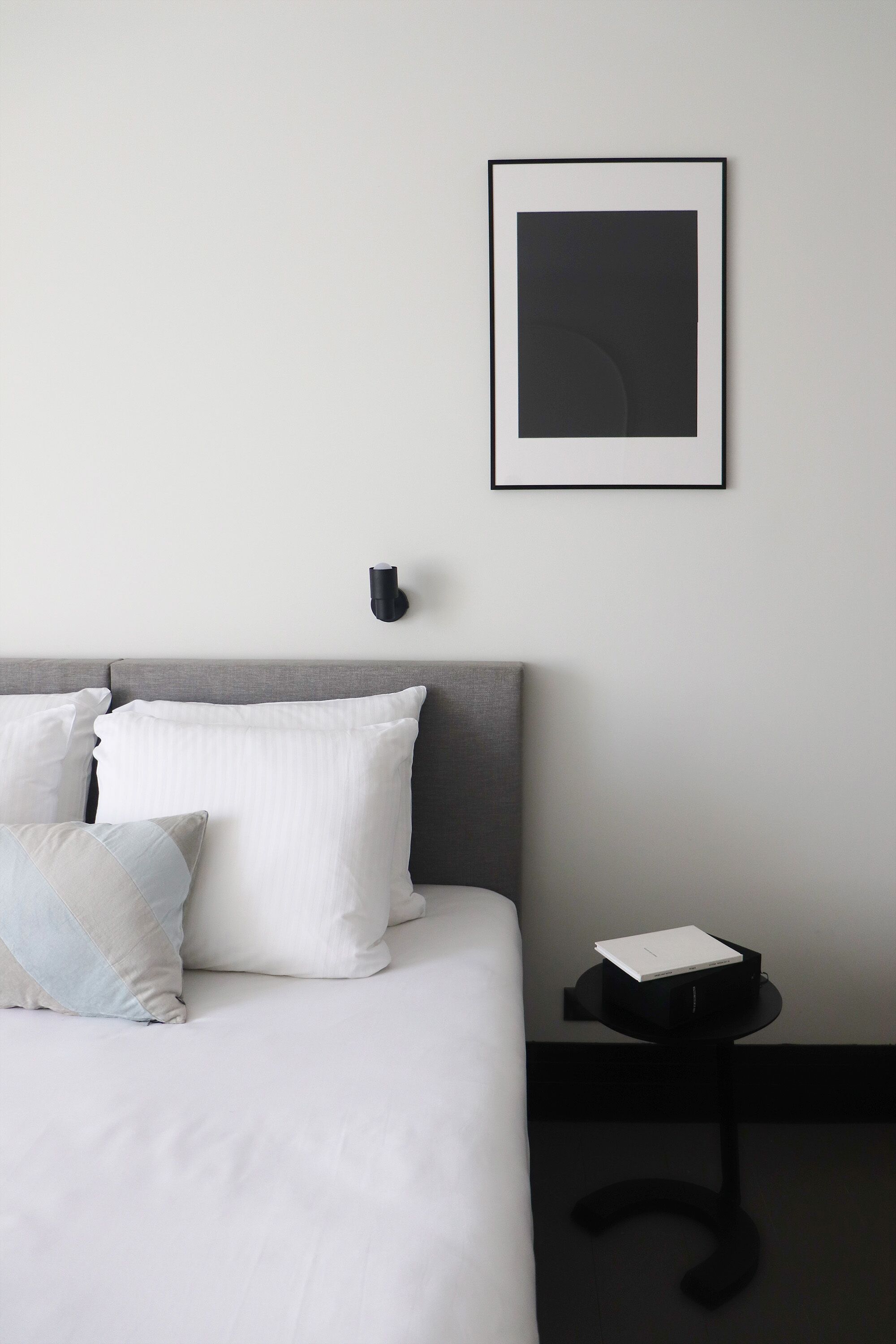 A neatly made bed with white linens and pillows, a black side table with a white object on it, and a framed picture on the wall.