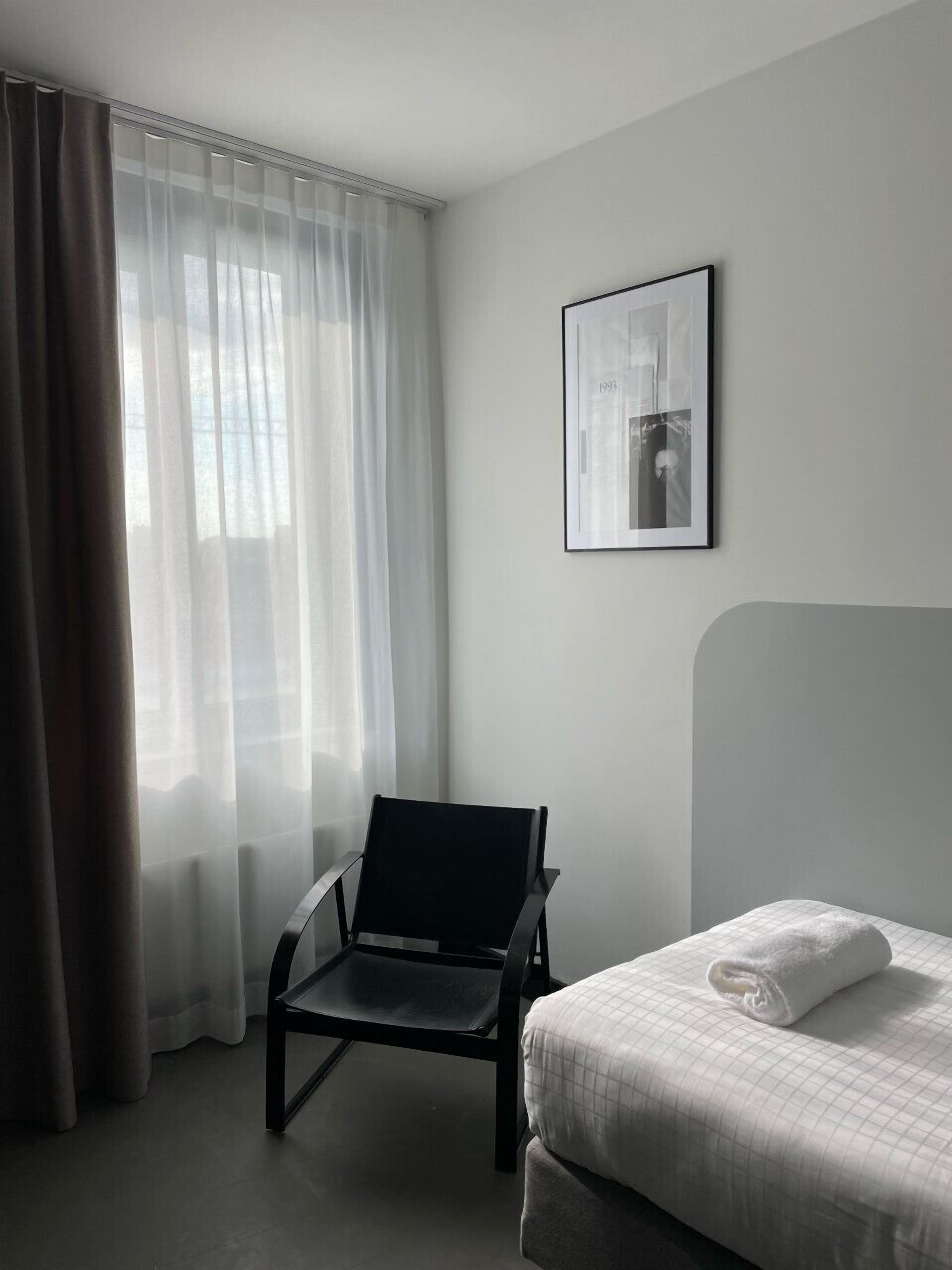 A hotel room with a bed, a chair, a window with sheer curtains, and a framed picture on the wall.