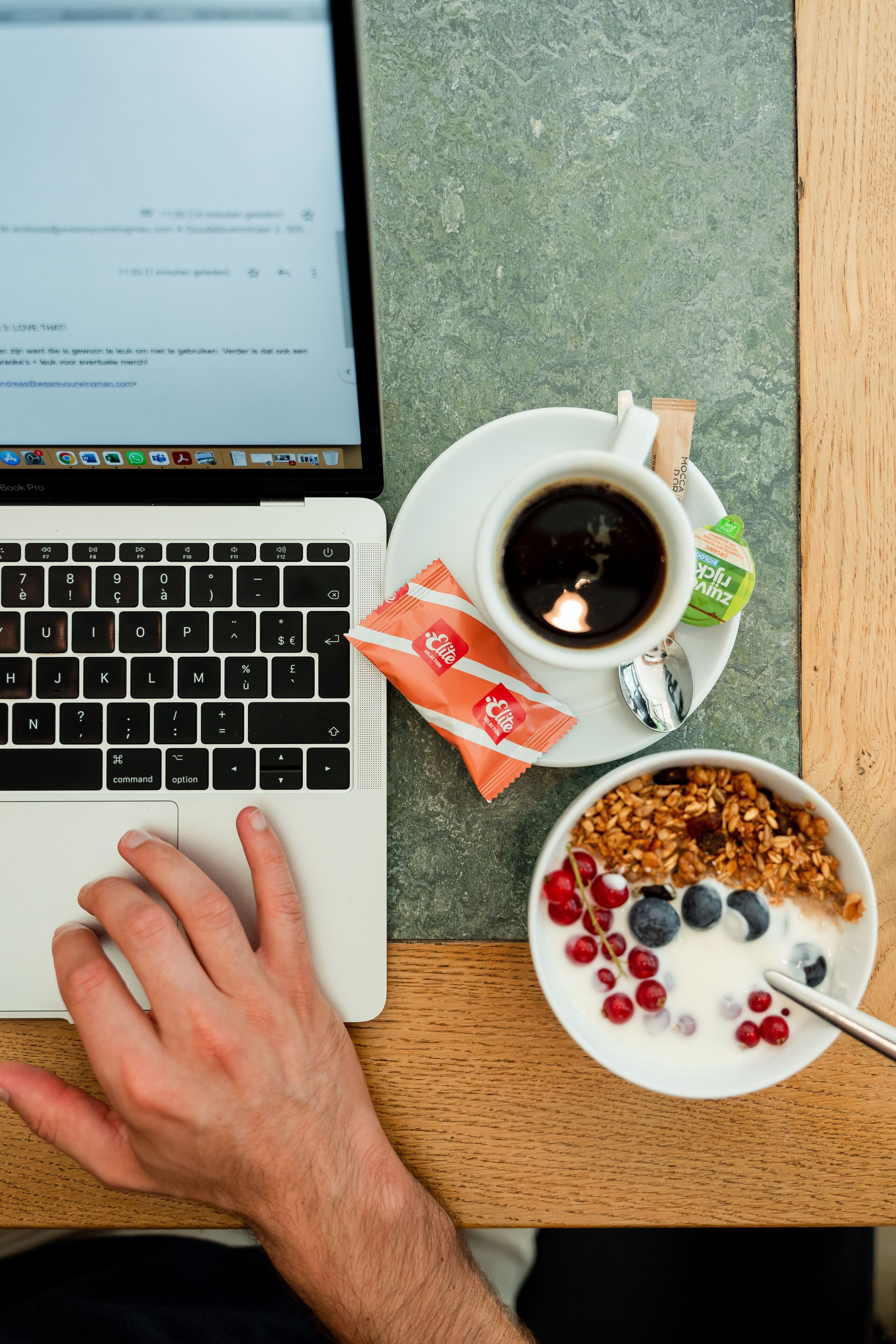 A laptop on a wooden table with a cup of coffee, a packet of snacks, and a bowl of cereal with fruits.