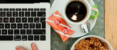 A laptop on a wooden table with a cup of coffee, a packet of snacks, and a bowl of cereal with fruits.