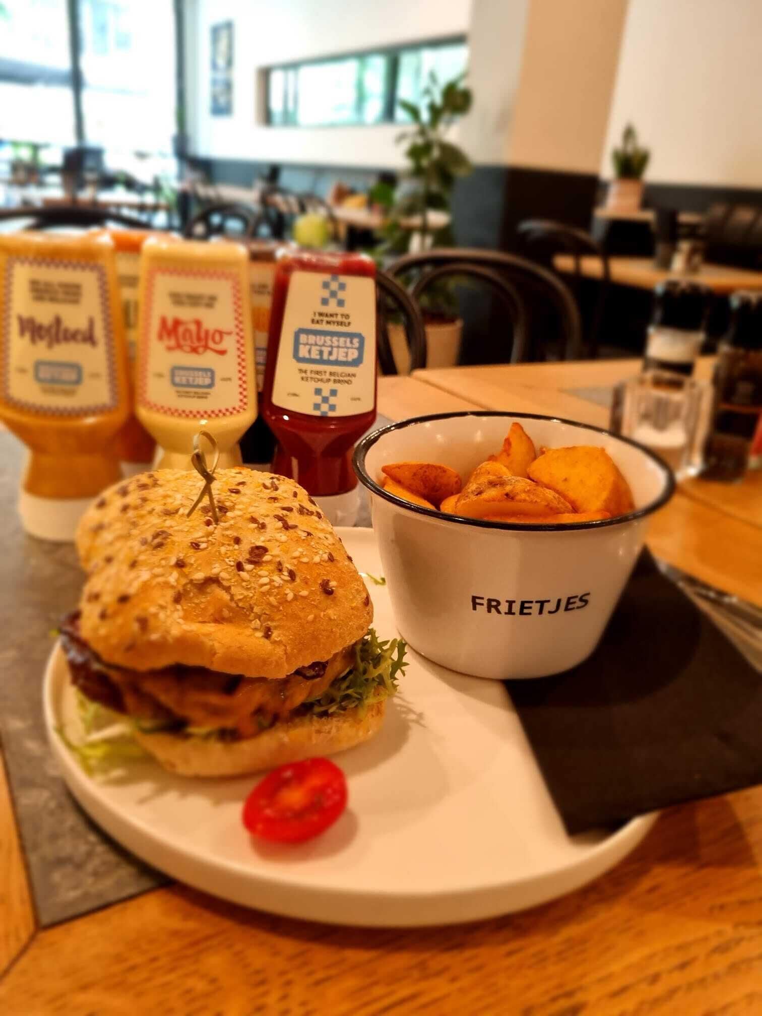 Un plato con una hamburguesa, papas fritas y kétchup, con condimentos etiquetados como 'Mayo' y 'Ketchup de Bruselas' sobre una mesa.