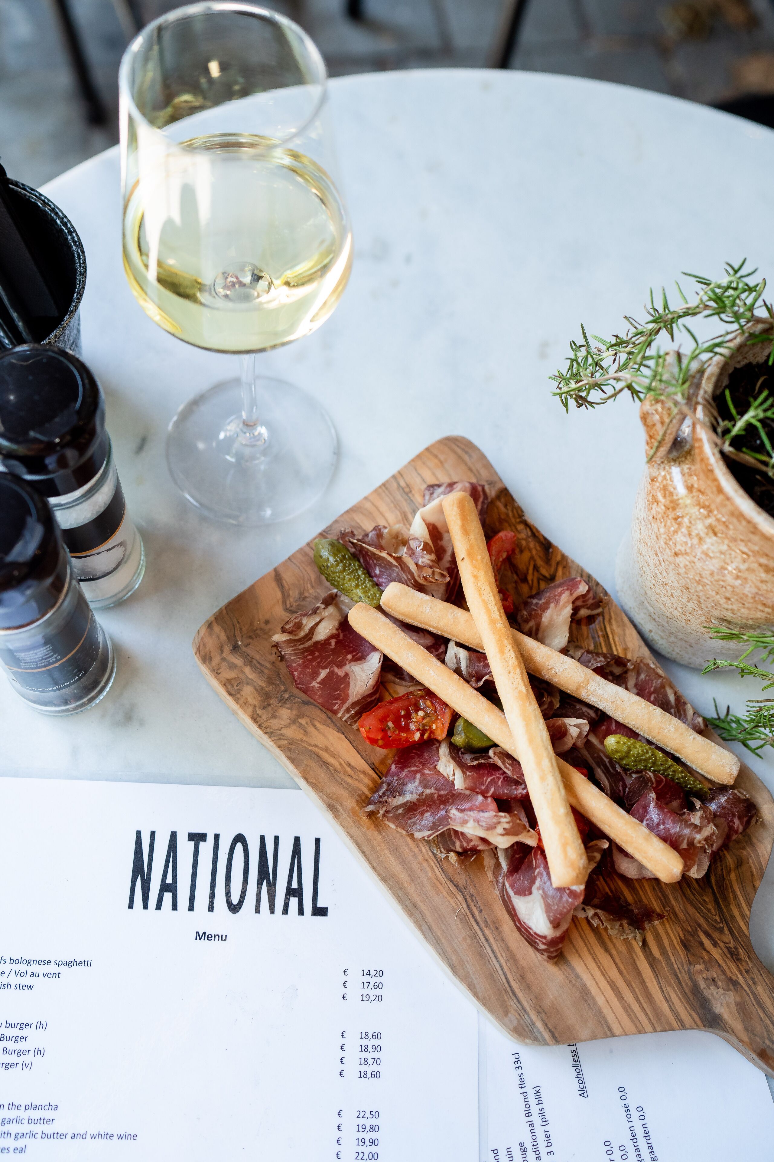 Eine Speisekarte mit verschiedenen Speisen und Preisen, ein Glas Weißwein sowie eine Holzplanke mit Fleisch und Gemüse.