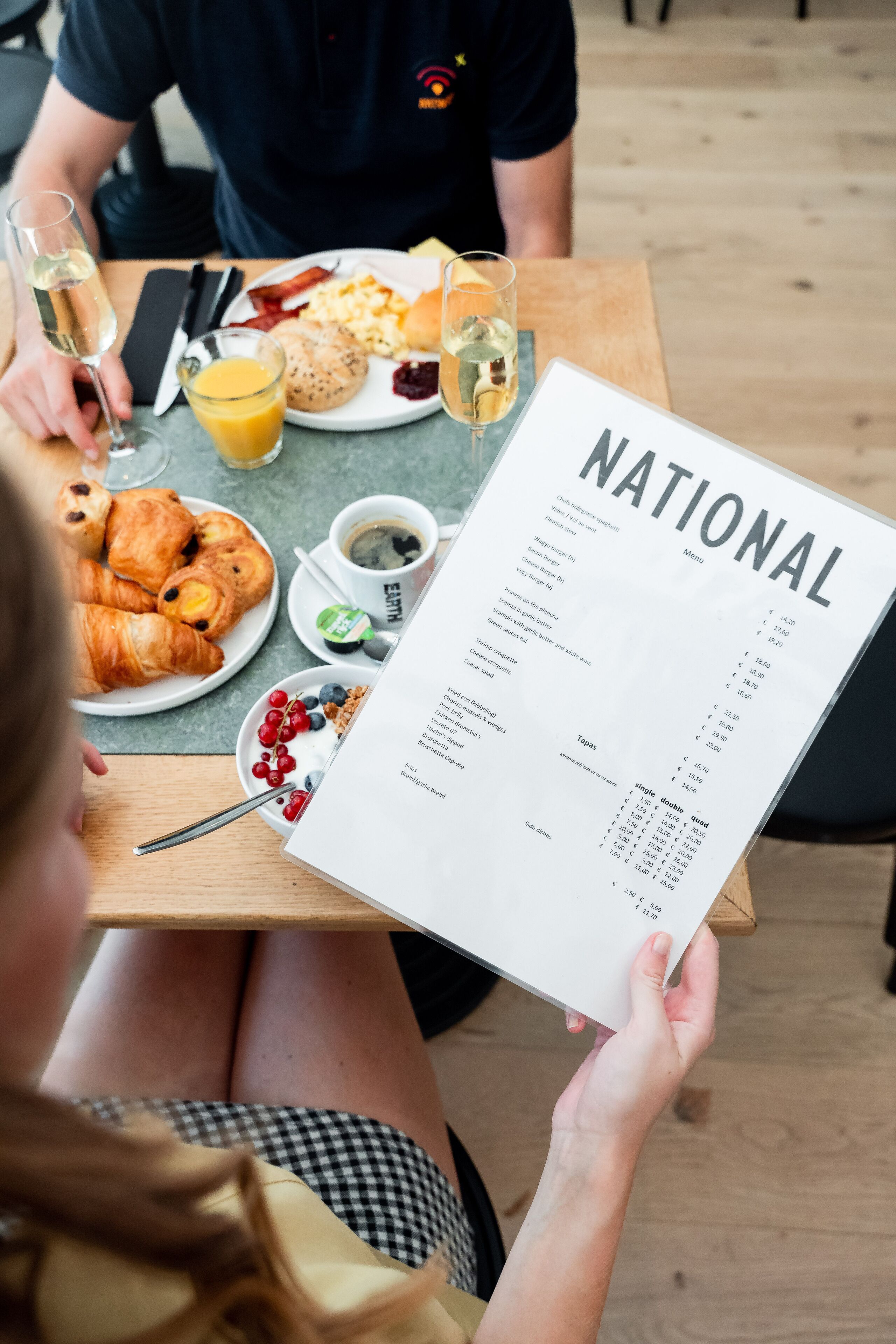A person holding a menu at a table with breakfast items like croissants, eggs, and juice.