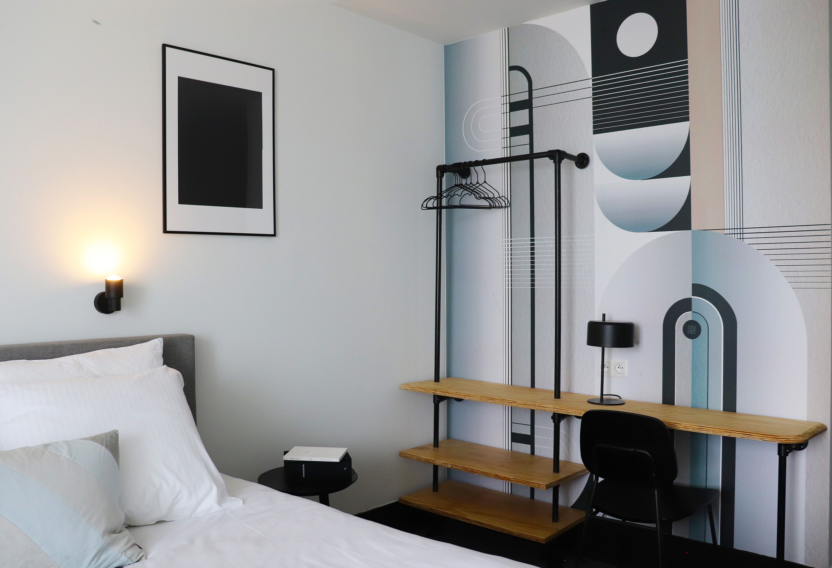 Une chambre d’hôtel moderne équipée d’un lit, d’un bureau avec une chaise, d’un porte-manteau mural et d’un mur orné d’œuvres d’art abstraites.