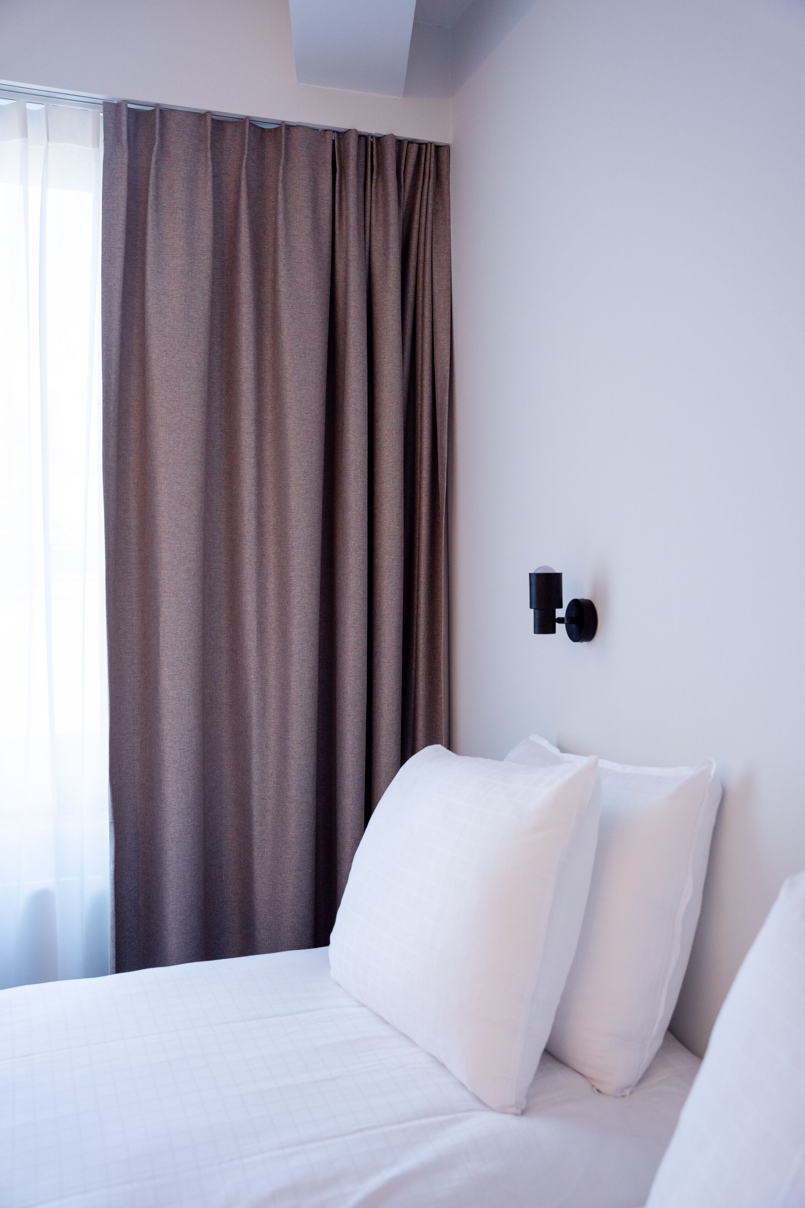 A hotel room with a bed, white bedding, a brown curtain, and a wall-mounted light fixture.