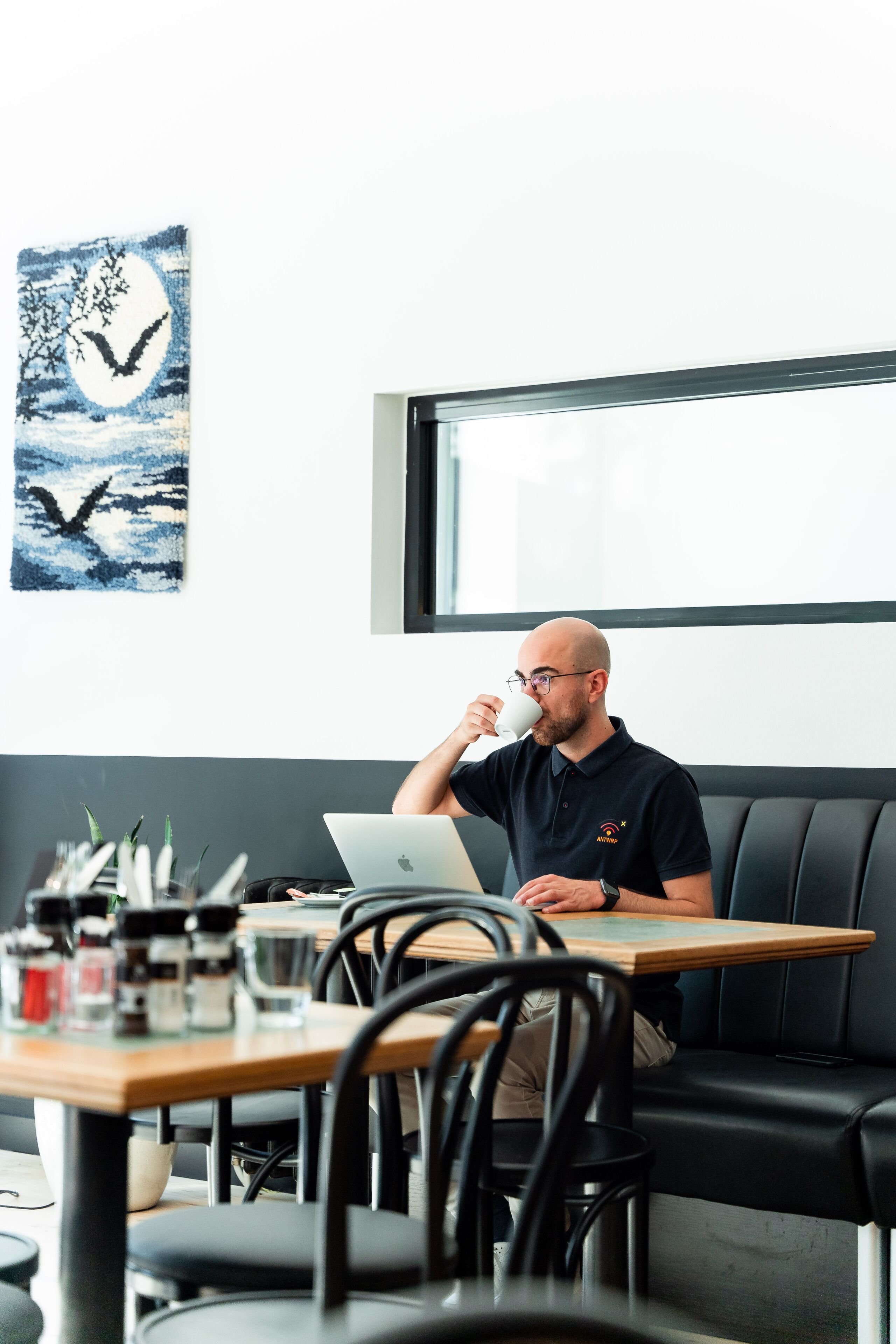 A man sitting at a table with a laptop, a window in the background, and artwork on the wall.