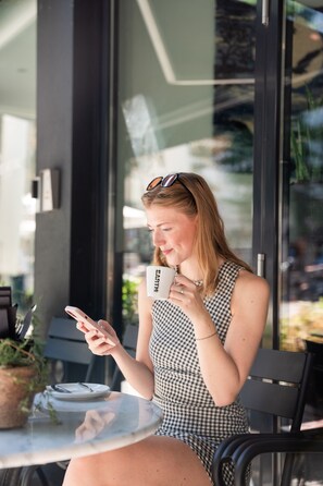 Una persona sentada a una mesa con una taza y un teléfono inteligente, vestida con un vestido a cuadros, con un ventanal de vidrio al fondo.
