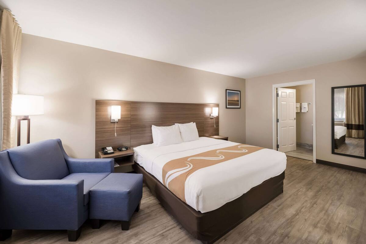 Quarto de hotel com cama, escrivaninha, cadeira e mesa de cabeceira.