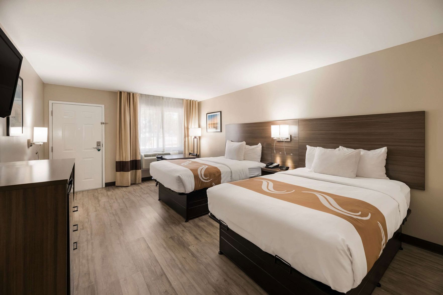 A hotel room with two beds, a desk, a chair, a wardrobe, and a window with curtains.