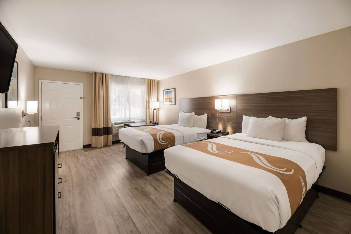 Quarto de hotel com duas camas, uma escrivaninha, uma cadeira, um guarda-roupa e uma janela com cortinas.