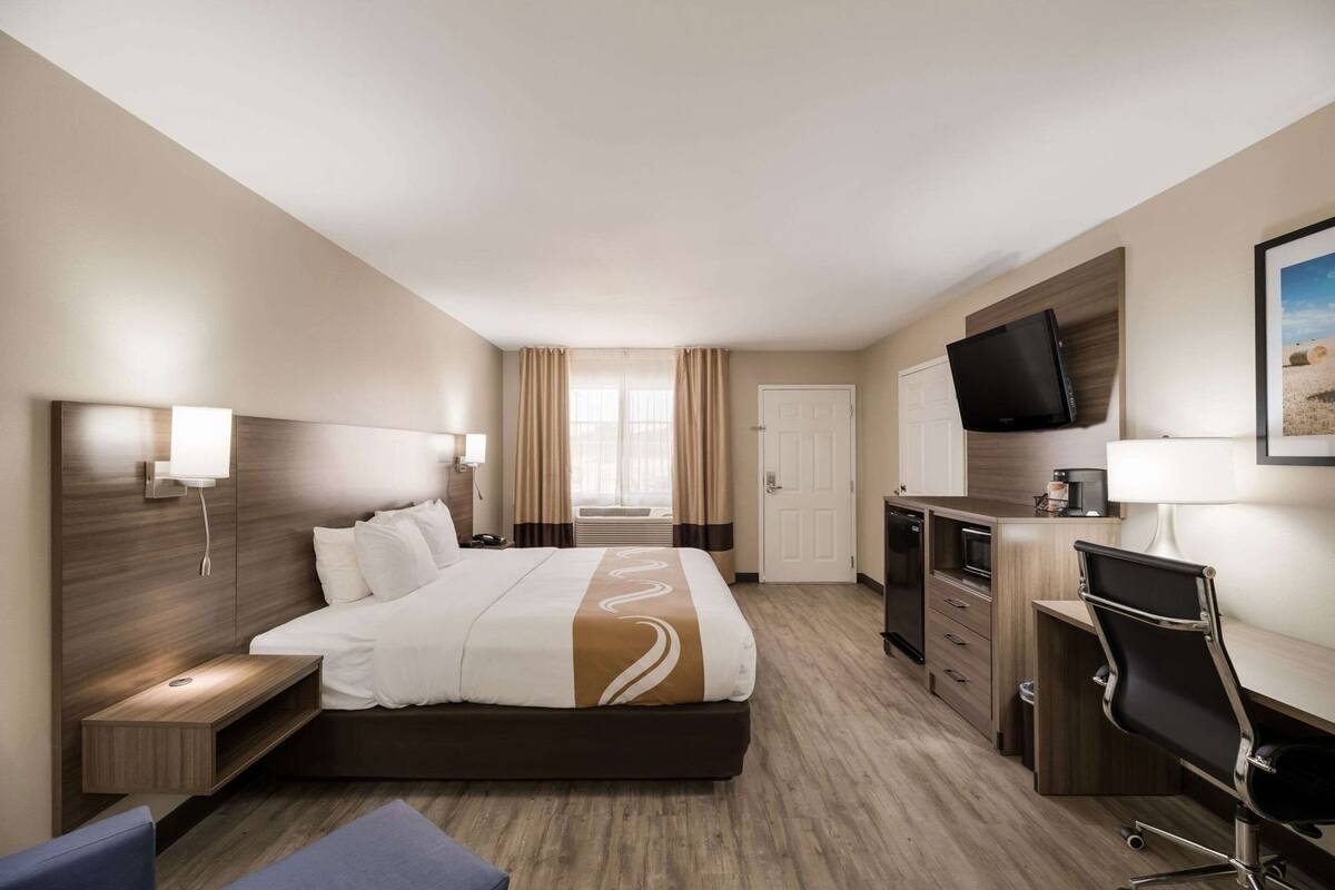 Quarto de hotel com cama, mesas de cabeceira, escrivaninha, cadeira, televisão e janela com cortinas.