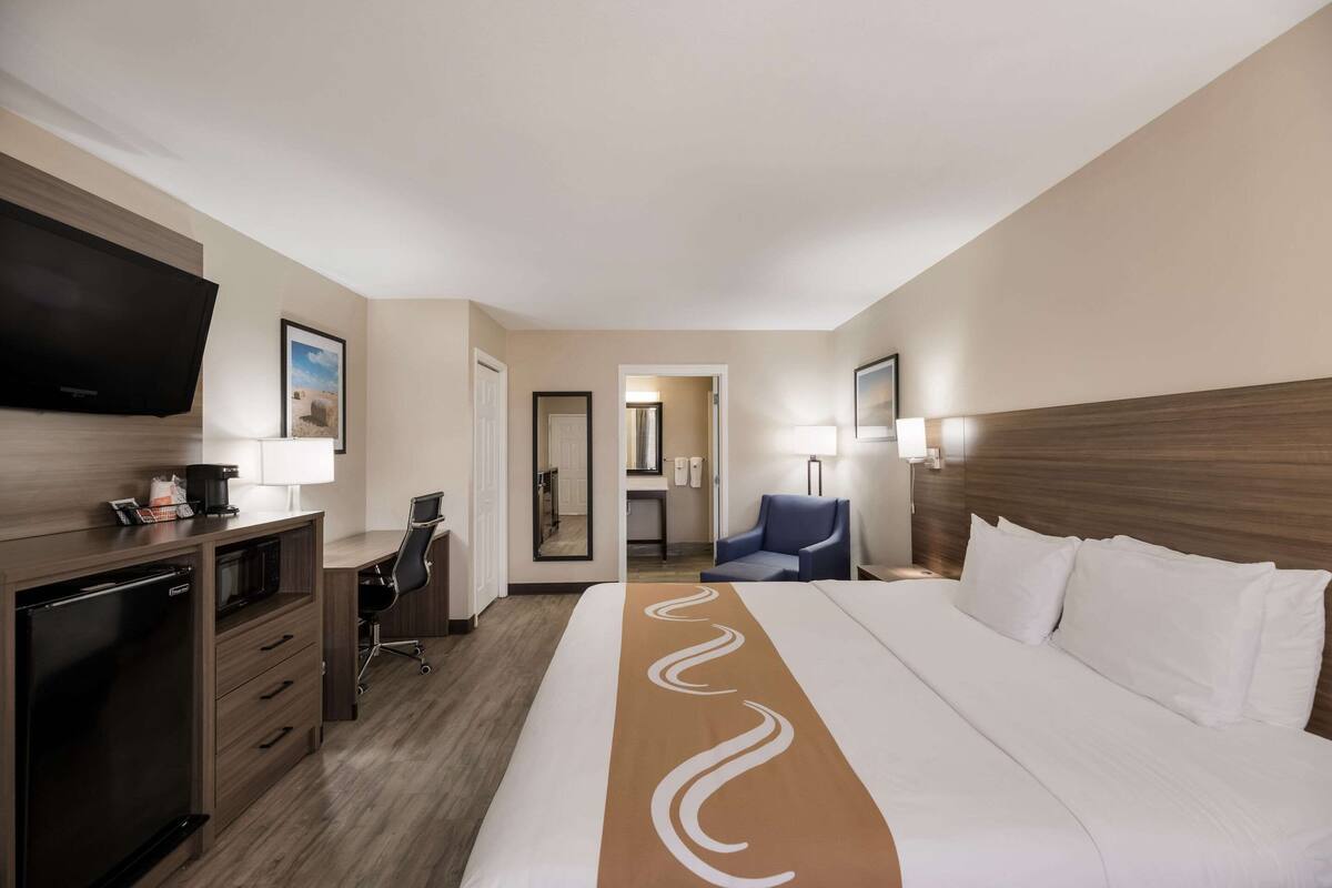 Quarto de hotel com cama, escrivaninha com computador, televisão e espelho.