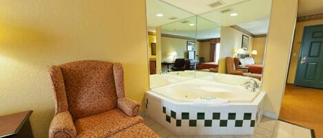 A hotel room with a large bathtub, a patterned armchair, and a mirror reflecting the room.