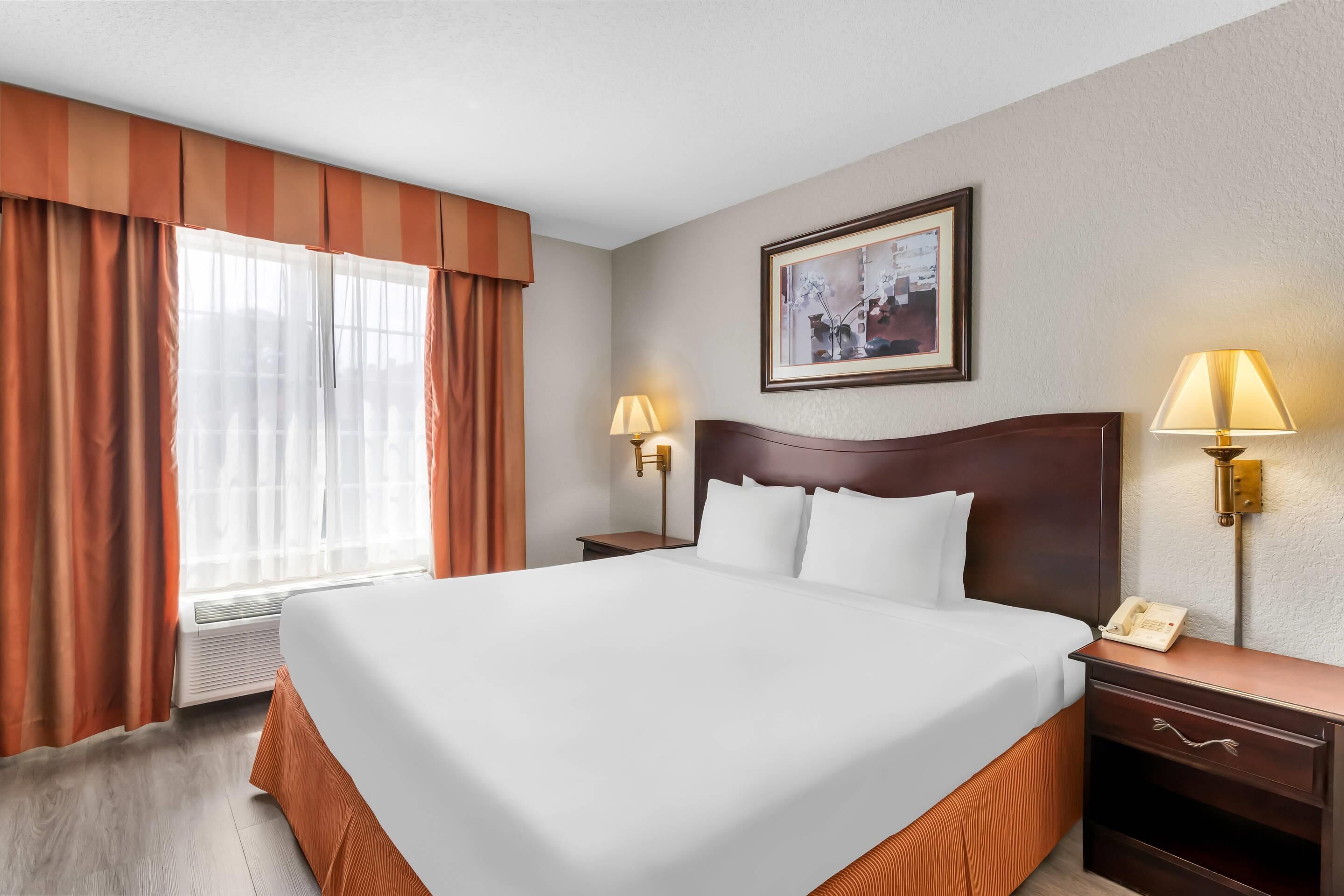 Una habitación de hotel con una cama grande, dos mesitas de noche con lámparas, un cuadro enmarcado en la pared y una ventana con cortinas.