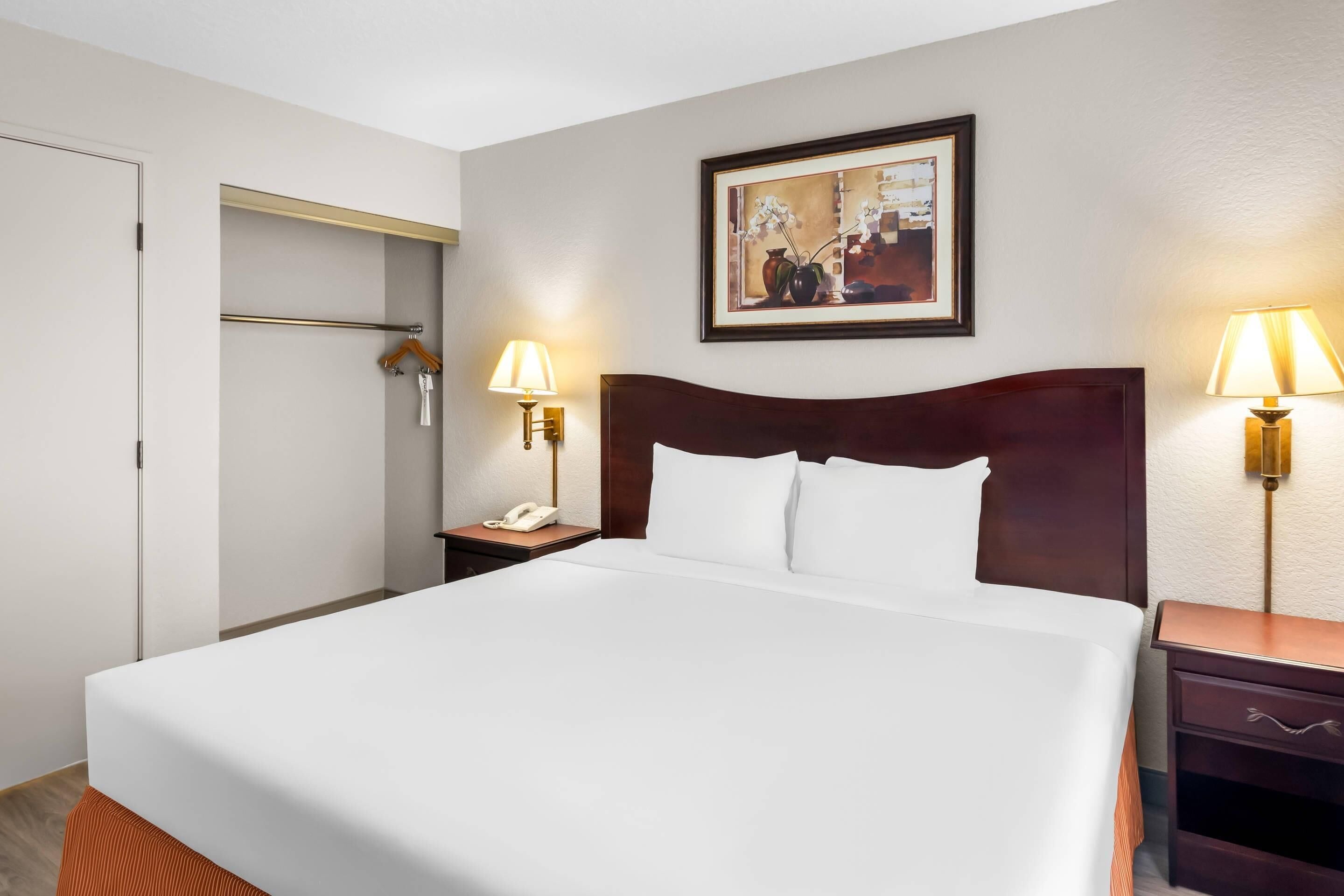 Una habitación de hotel con una cama, lámparas de noche, una mesita de noche, un cuadro en la pared y una puerta.