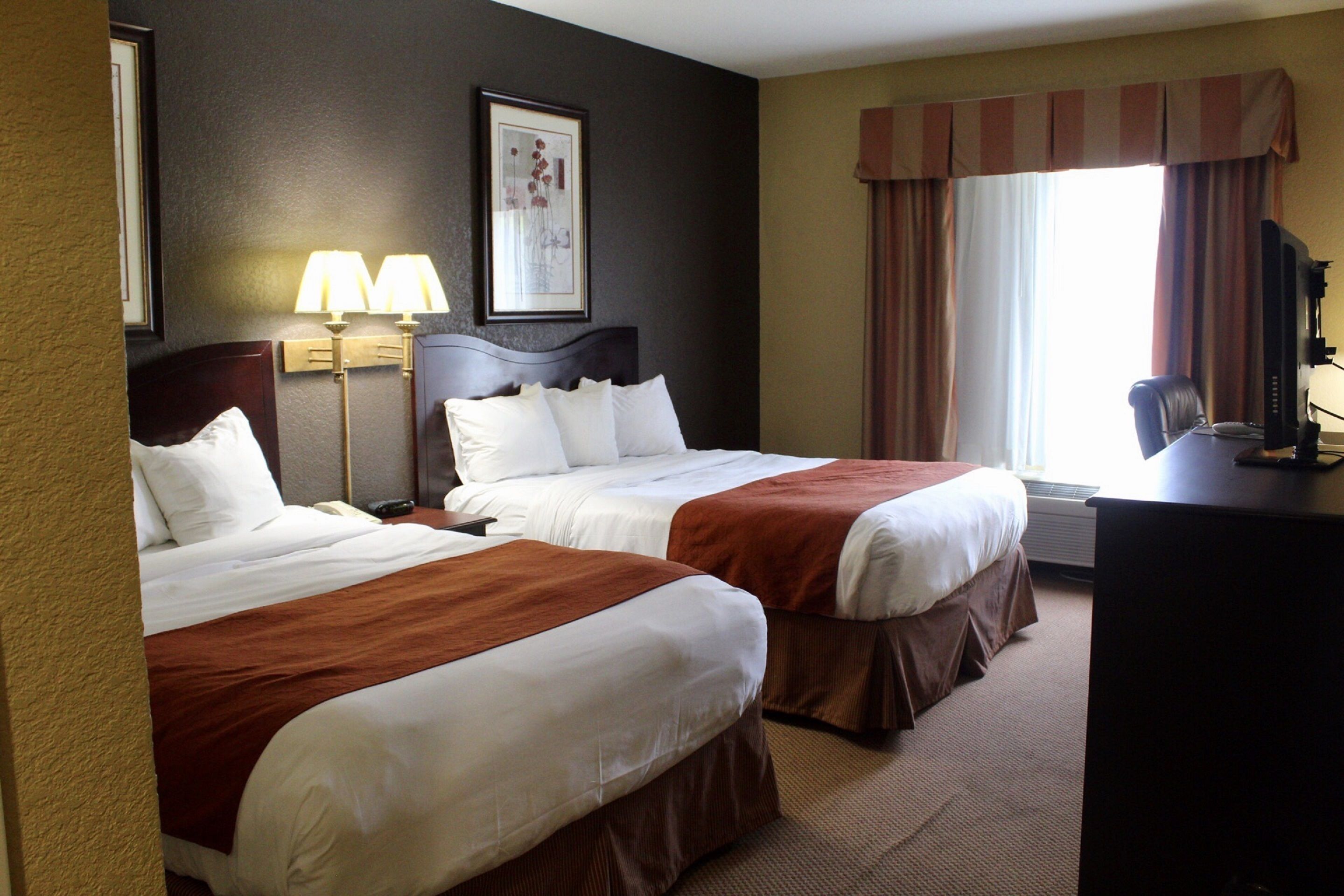 A hotel room with two beds, a desk, a chair, a lamp, and a window with curtains.