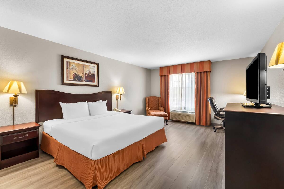 A hotel room with a large bed, two bedside tables with lamps, a desk with a TV, a chair, and a window with curtains.