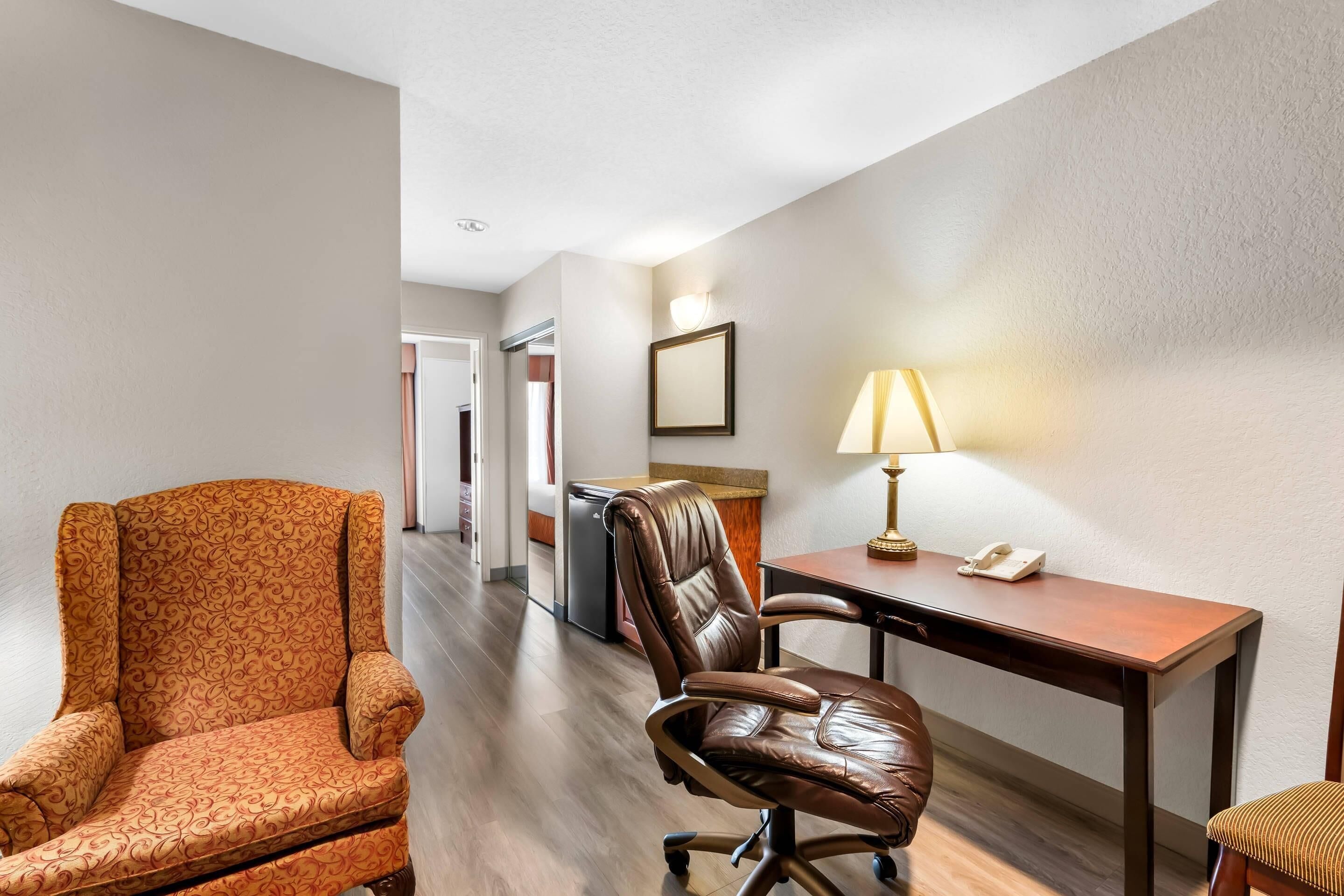 A hotel room with a patterned armchair, a leather office chair, a desk with a lamp, and a telephone.