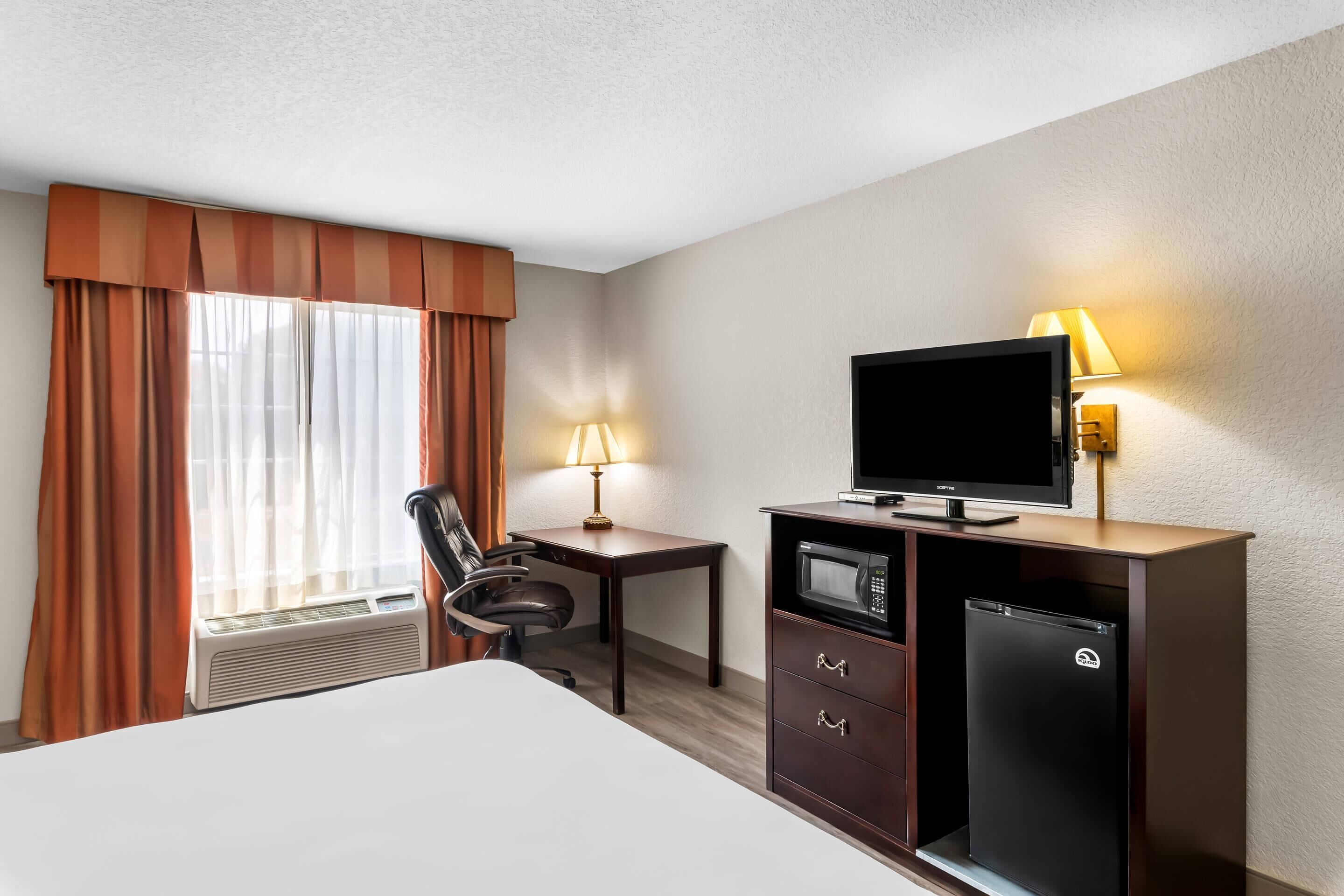 A hotel room with a bed, a desk with a lamp, a TV, a mini-fridge, and a chair.