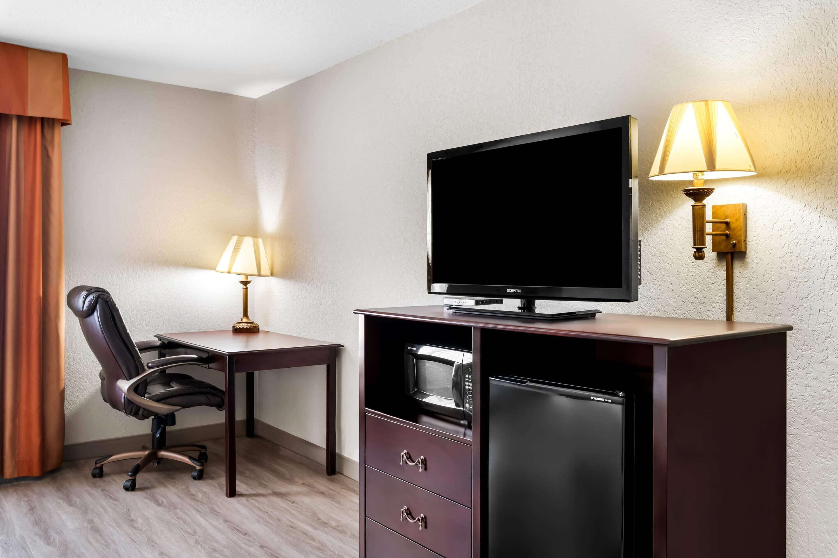 Habitación de hotel con televisión de pantalla plana, escritorio con lámpara, silla, microondas y refrigerador.
