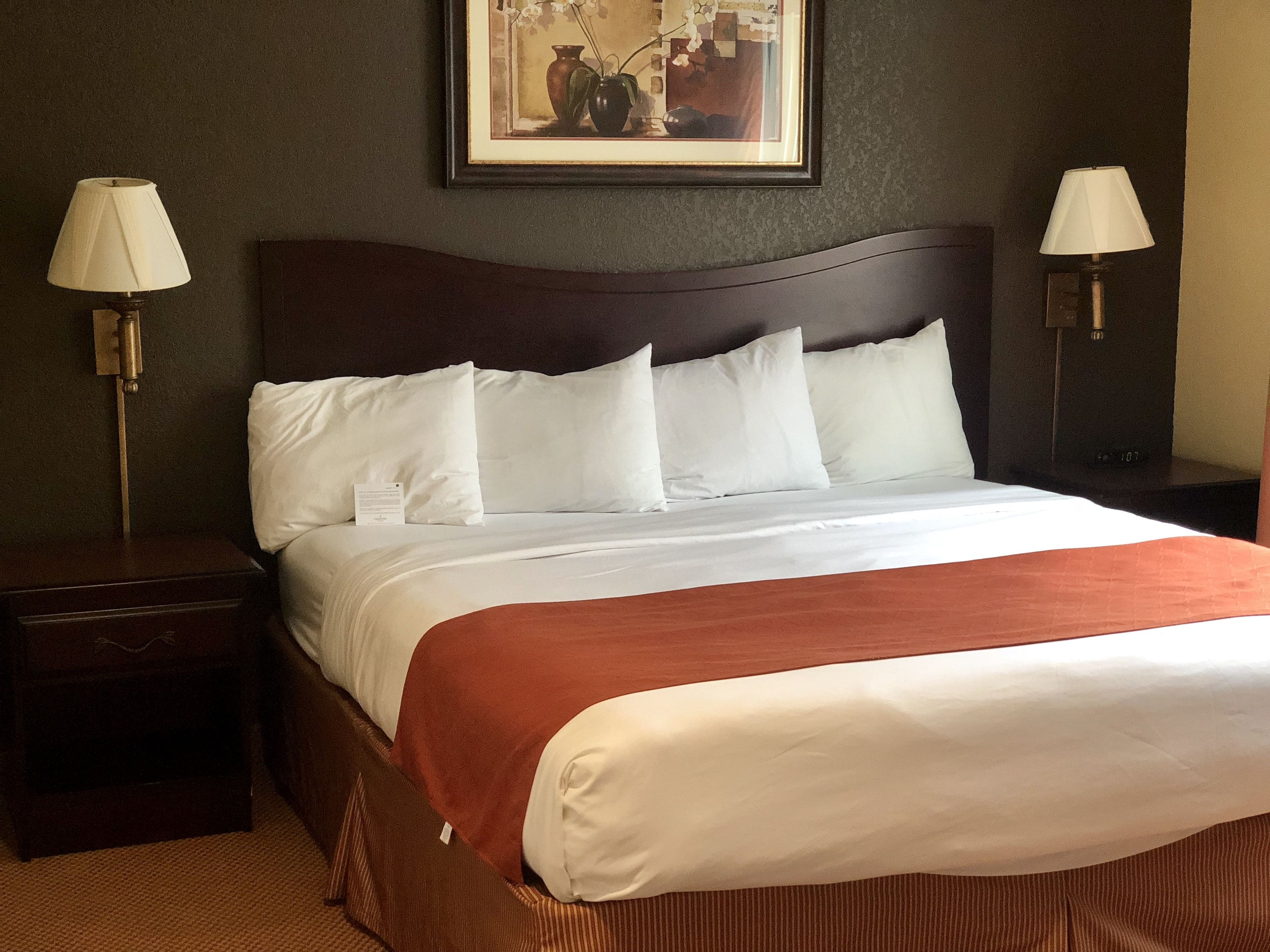 Una cama bien hecha con sábanas blancas y un plaid marrón, con dos mesitas de noche y lámparas a cada lado, en una habitación con una obra enmarcada sobre la cama.