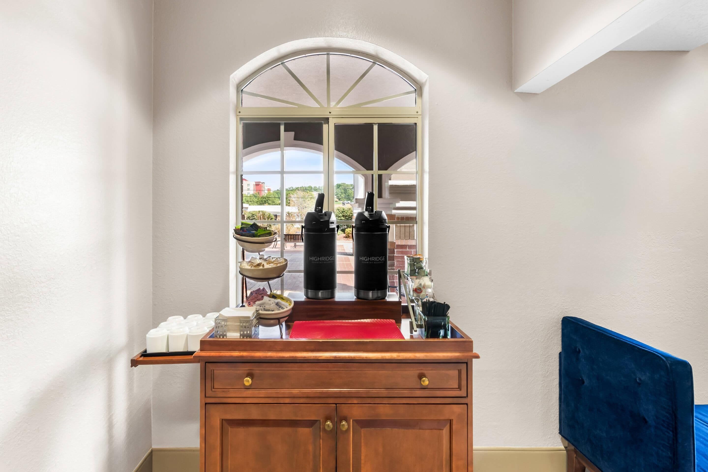 A hotel room with a wooden cabinet, a red tray, and two black water bottles labeled 'HIGHRIDGE' on a window sill.