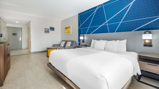 Ein modernes Hotelzimmer mit einem großen Bett, einem grauen Sofa, einem Holfnachtkästchen und einem blauen, geometrischen Wanddesign.