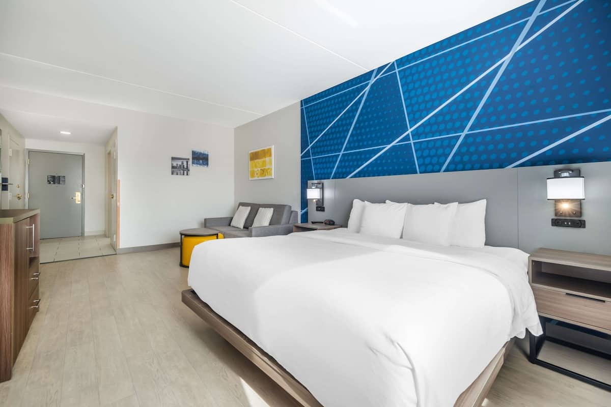 Ein modernes Hotelzimmer mit einem großen Bett, einem grauen Sofa, einem Holfnachtkästchen und einem blauen, geometrischen Wanddesign.