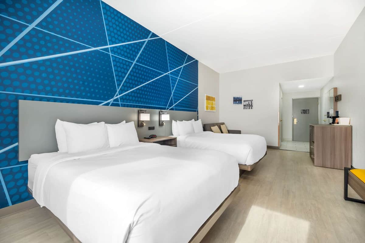 Ein Hotelzimmer mit zwei Betten, einer blauen Wand mit geometrischem Muster, einem Holzschrank und einem Spiegel.
