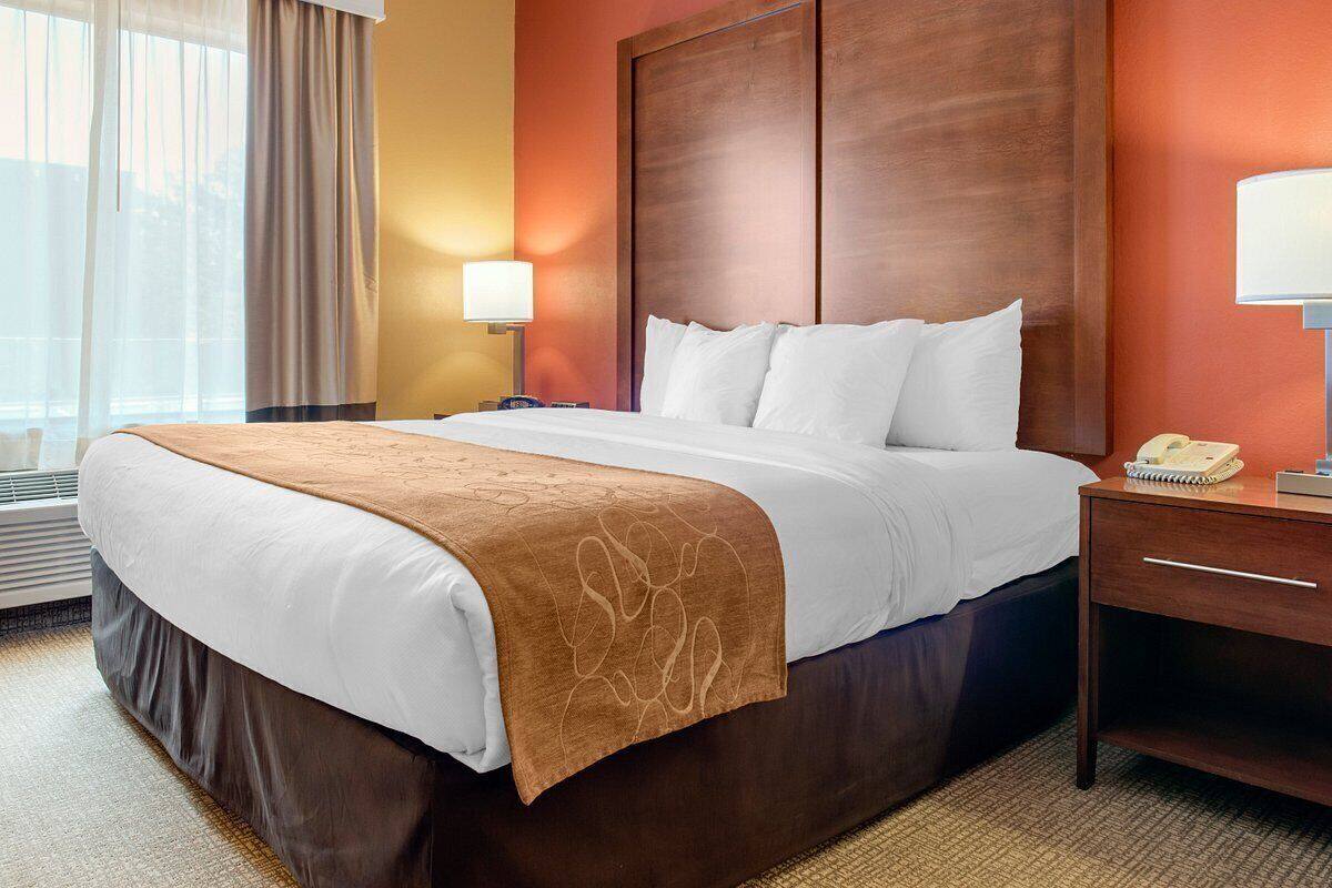 Une chambre d’hôtel avec un grand lit, une tête de lit en bois, une table de chevet avec une lampe et une fenêtre avec des rideaux.