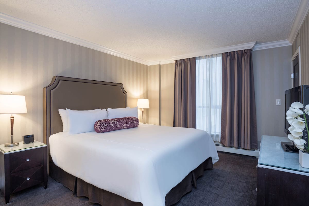 A hotel room with a large bed, two bedside lamps, a nightstand, a dresser, and a large window with curtains.