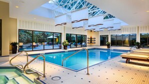 Una piscina interior con paredes de vidrio transparente, tragaluces y una terraza de madera con sillones de descanso.