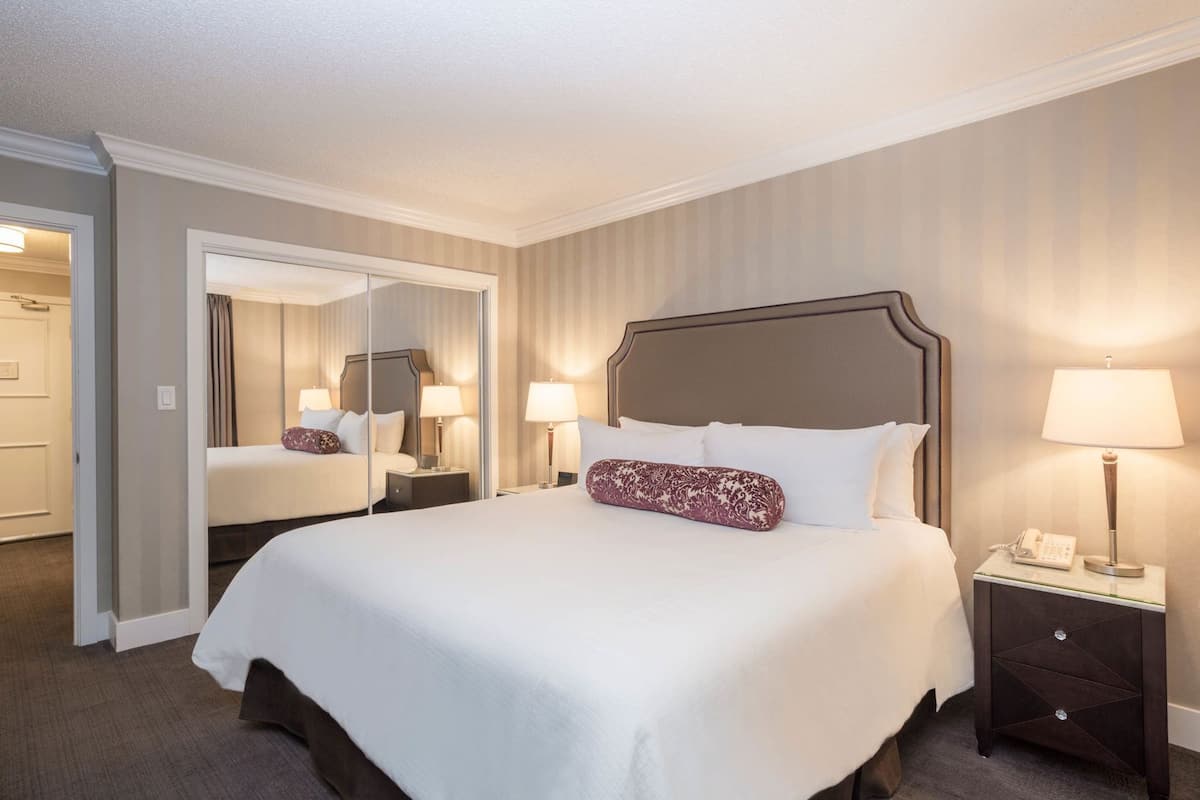 A hotel room with a large bed, bedside lamps, a nightstand, a mirror, and a door leading to another room.