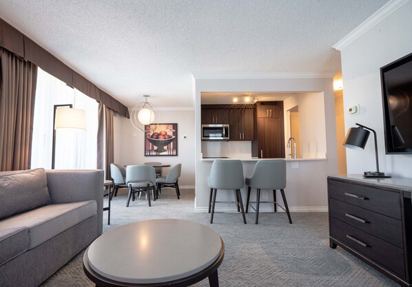A modern living room with a sofa, a round coffee table, and a dining area with chairs and a table. There is a kitchen area with a microwave, cabinets, and a bar counter. - La Grande Residence Vancouver at The Sutton Place Hotel (Vancouver)