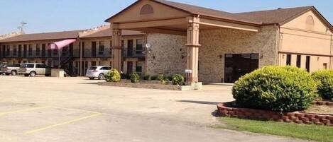 A motel with a covered parking area, a stone facade, and a small landscaped area with bushes.