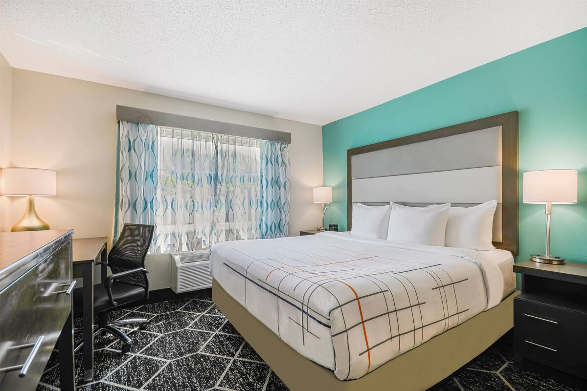 Une chambre d’hôtel comprenant un lit, deux lampes de chevet, un bureau avec une chaise, une fenêtre avec des rideaux et un climatiseur fixé au mur.