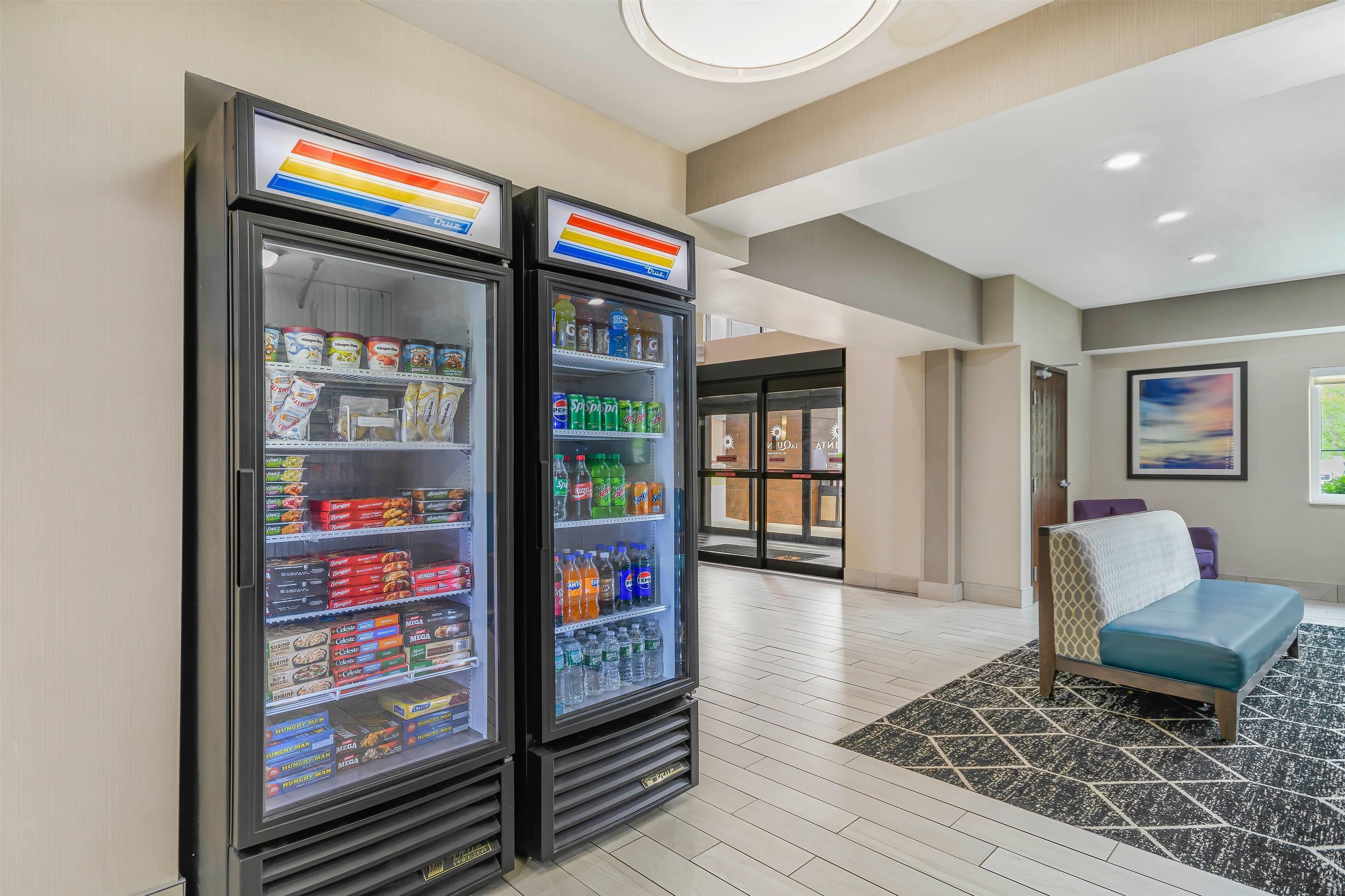 Two refrigerated beverage dispensers in a hotel lobby with a patterned rug and a bed in the background.
