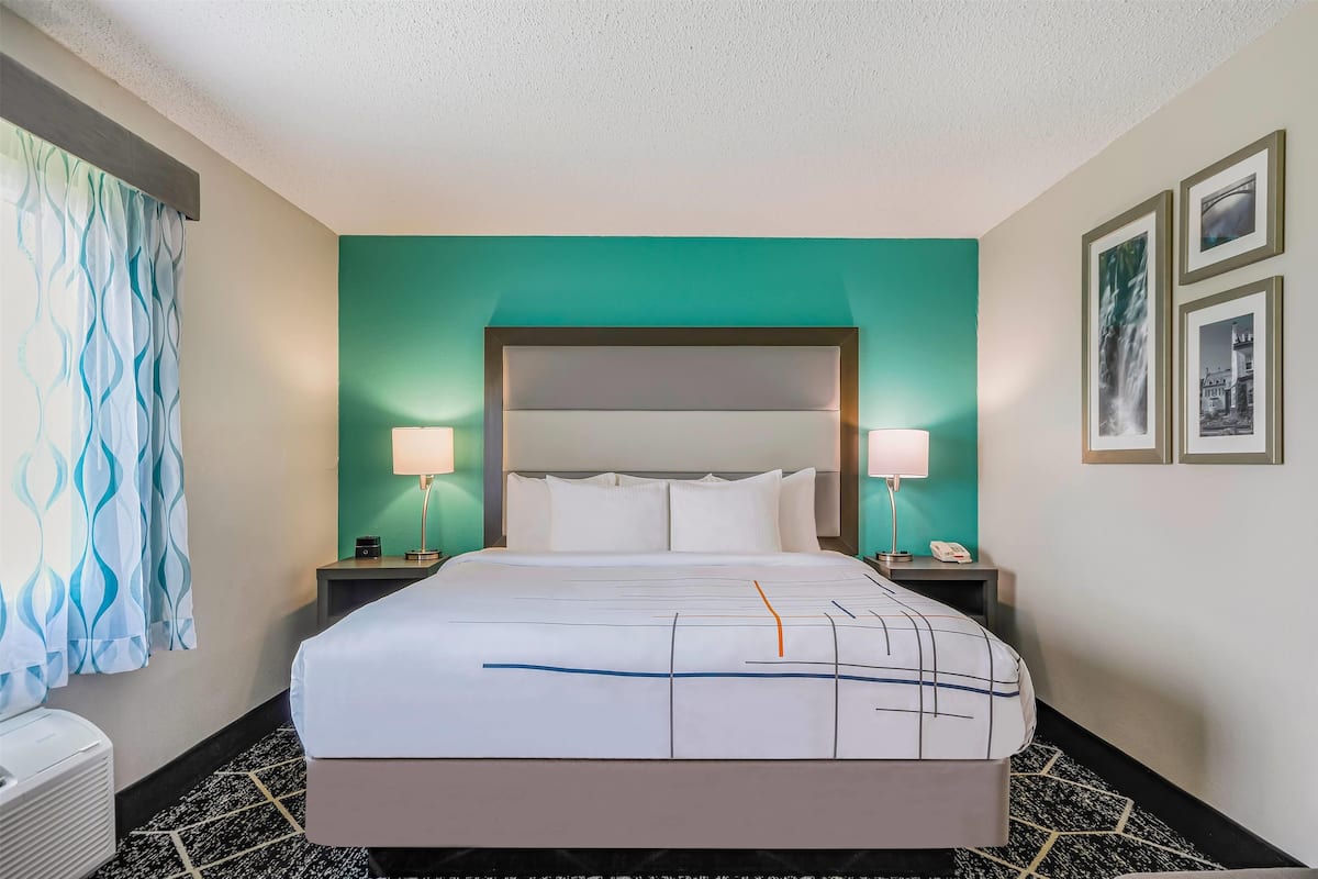 Une chambre d’hôtel comprenant un lit, deux tables de chevet, une tête de lit, une fenêtre avec des rideaux et un mur orné de tableaux.