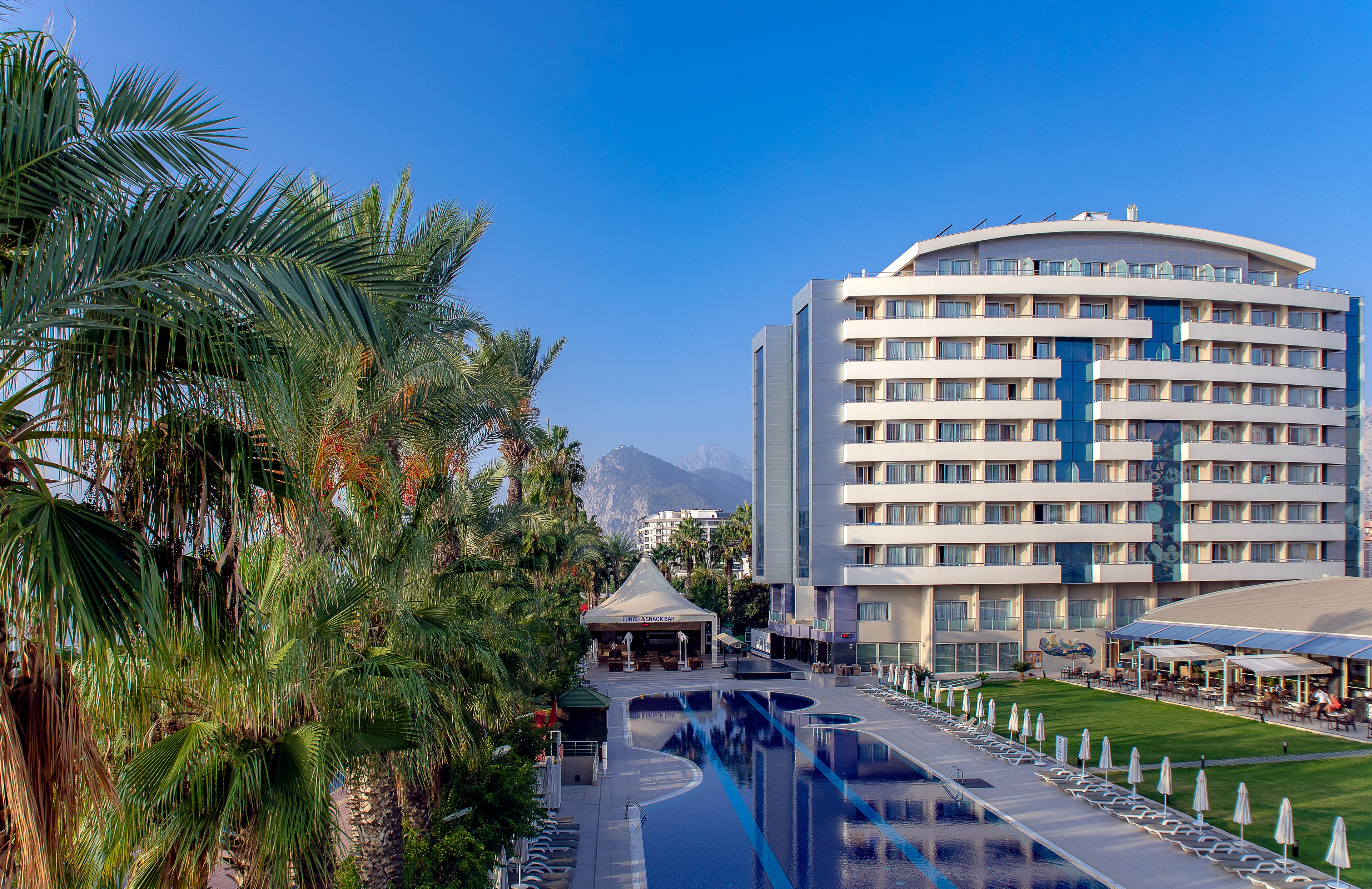 Een hotel met een groot zwembad, palmbomen en een bergachtig achtergrond.