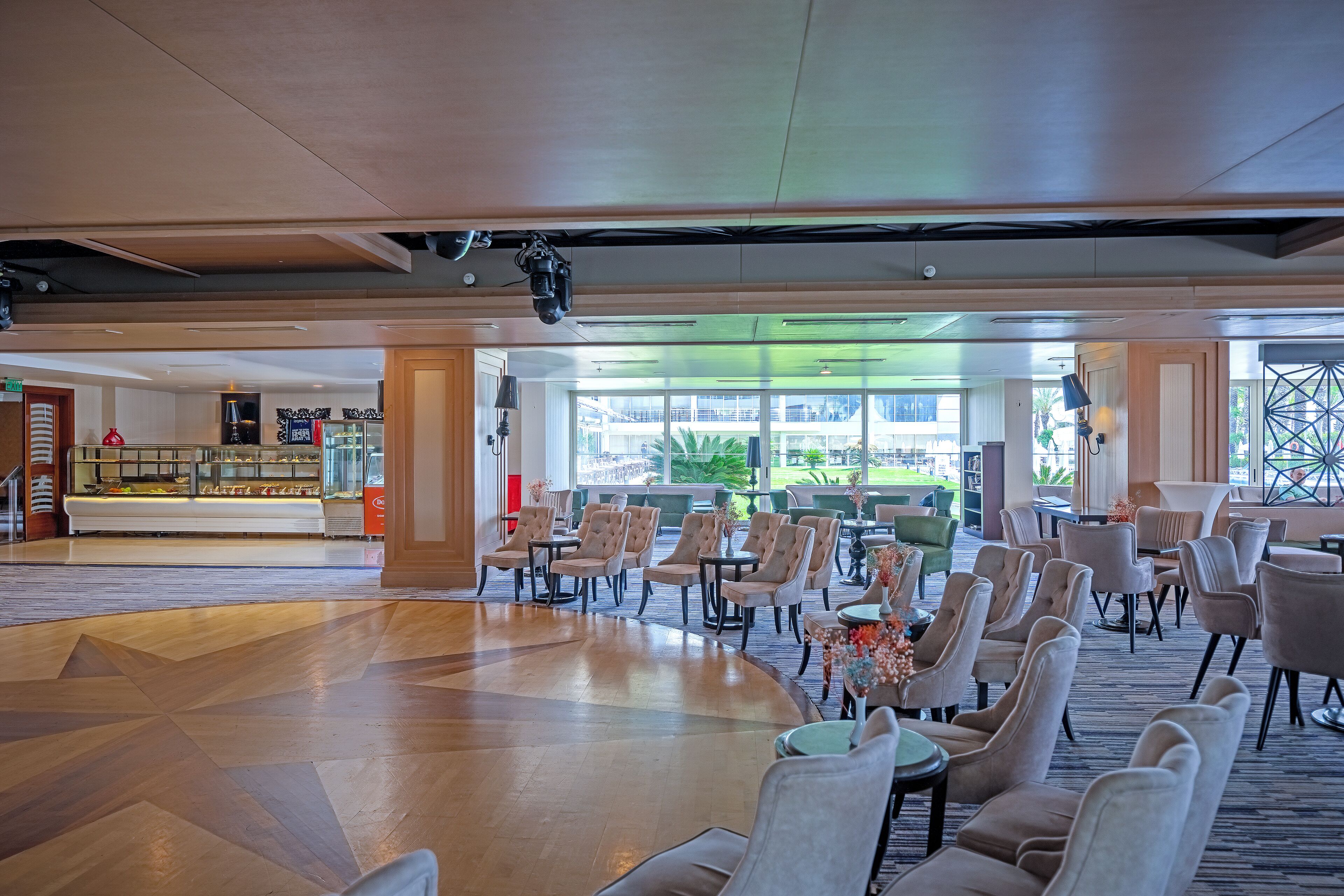 A spacious lobby with a modern design, featuring a central seating area with multiple chairs and tables, a food display counter, and large windows providing natural light and views of the outdoors.