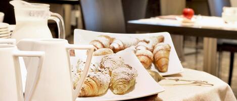 A breakfast setup with croissants and a pitcher of milk on a table, with chairs and a window in the background.
