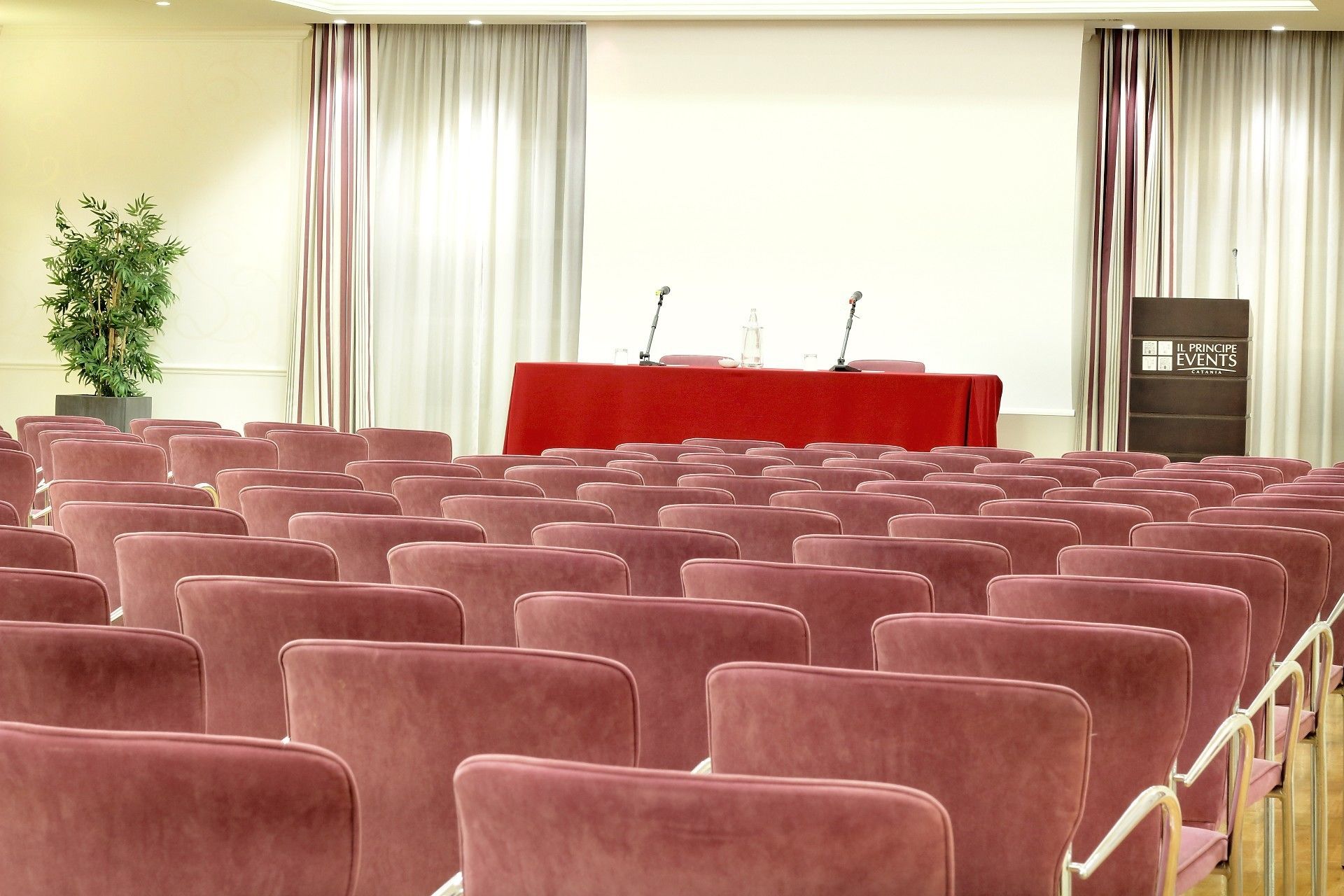 Un salón de conferencias con filas de sillas rojas, un atril y una pantalla para proyector.