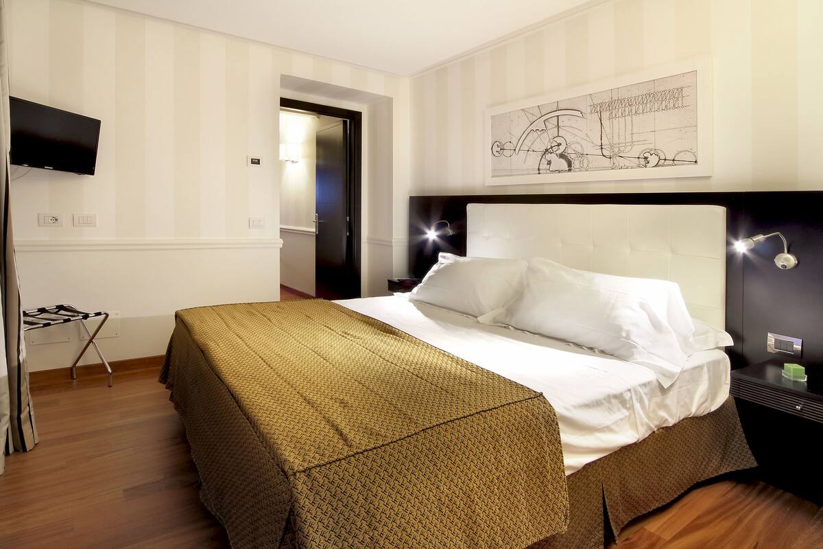 A hotel room with a bed, a TV mounted on the wall, a framed artwork above the bed, and a door leading to another room.