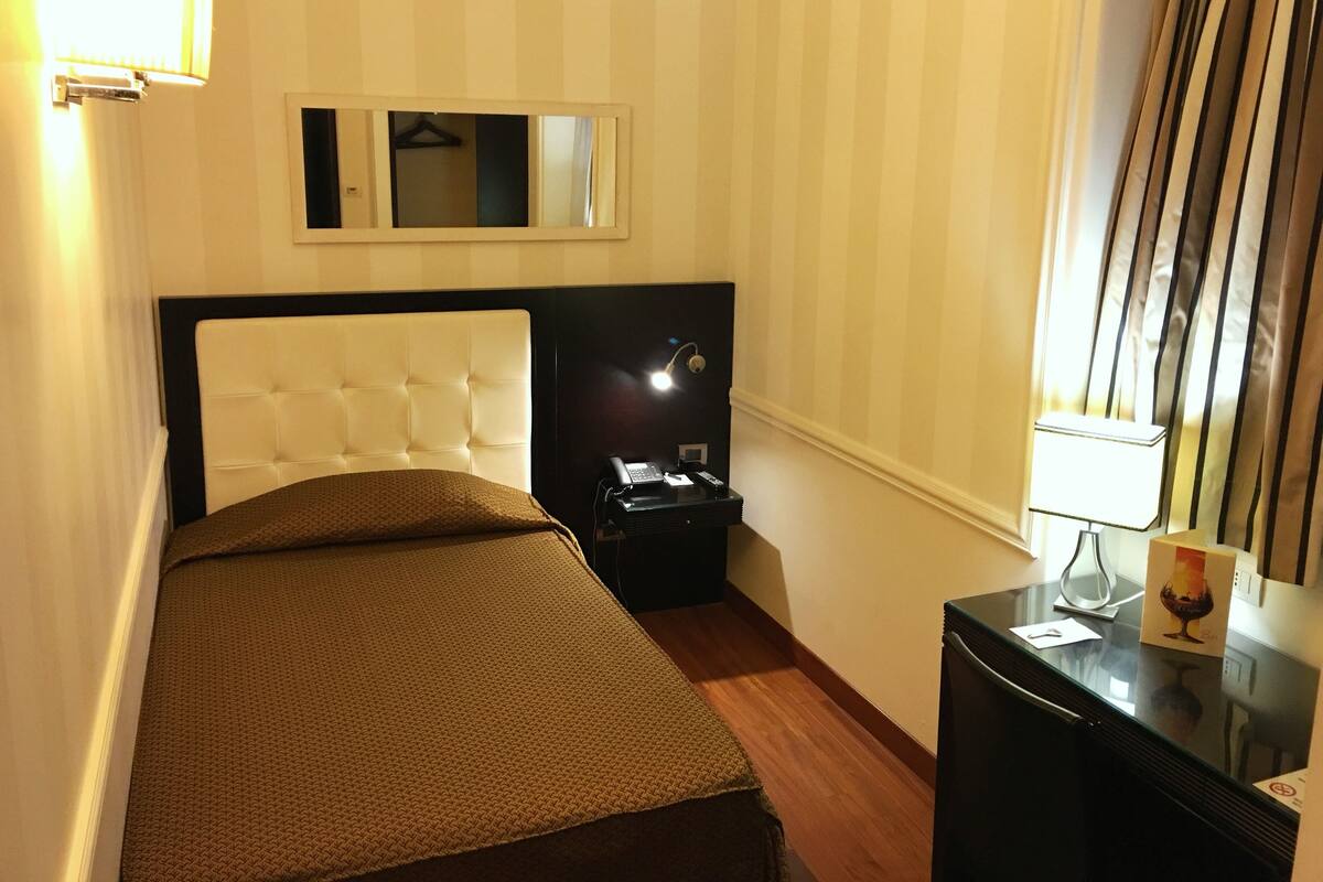A hotel room with a bed, a bedside table with a lamp, a mirror, and a window with curtains.